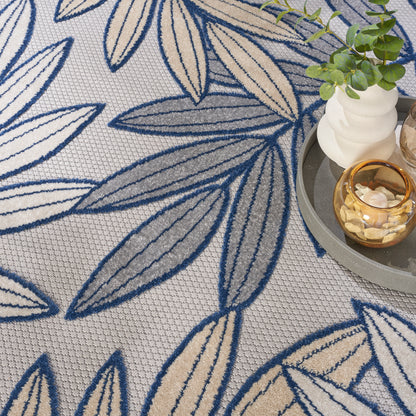 Beige rug with dark blue leaf pattern and textured weave; plant and candle on a gray round tray beside.