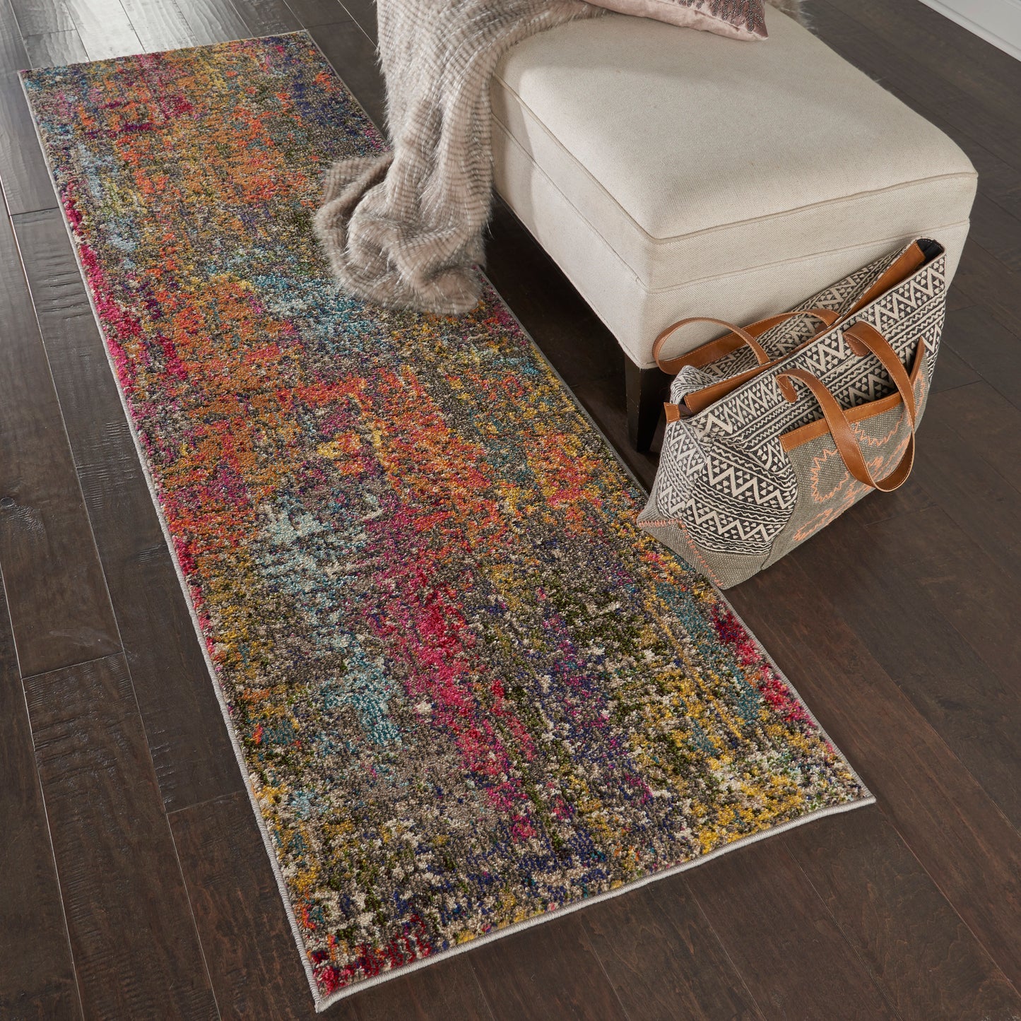 Colorful abstract runner rug on dark wood floor beside beige ottoman with knit throw and patterned tote.