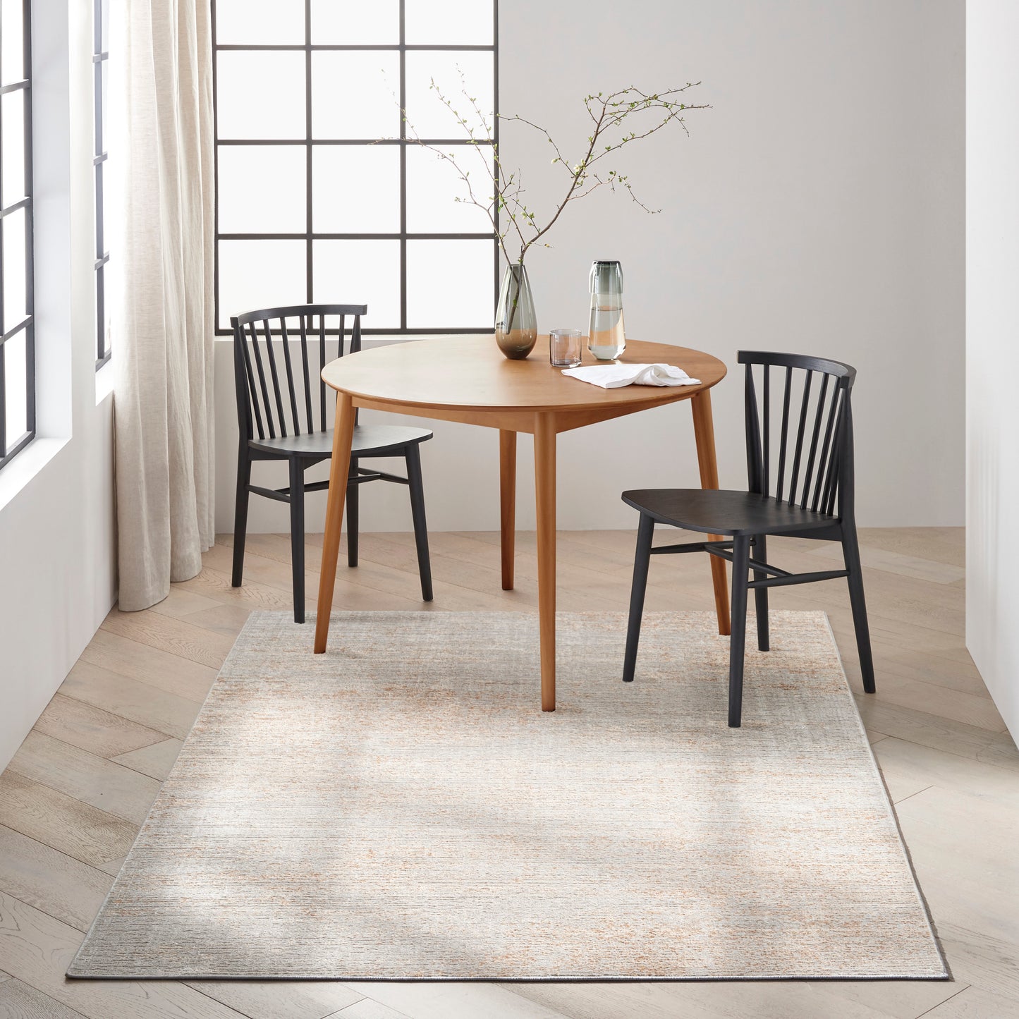 Round wooden dining table with two black chairs, glass pitcher and vase with branches on a beige rug.