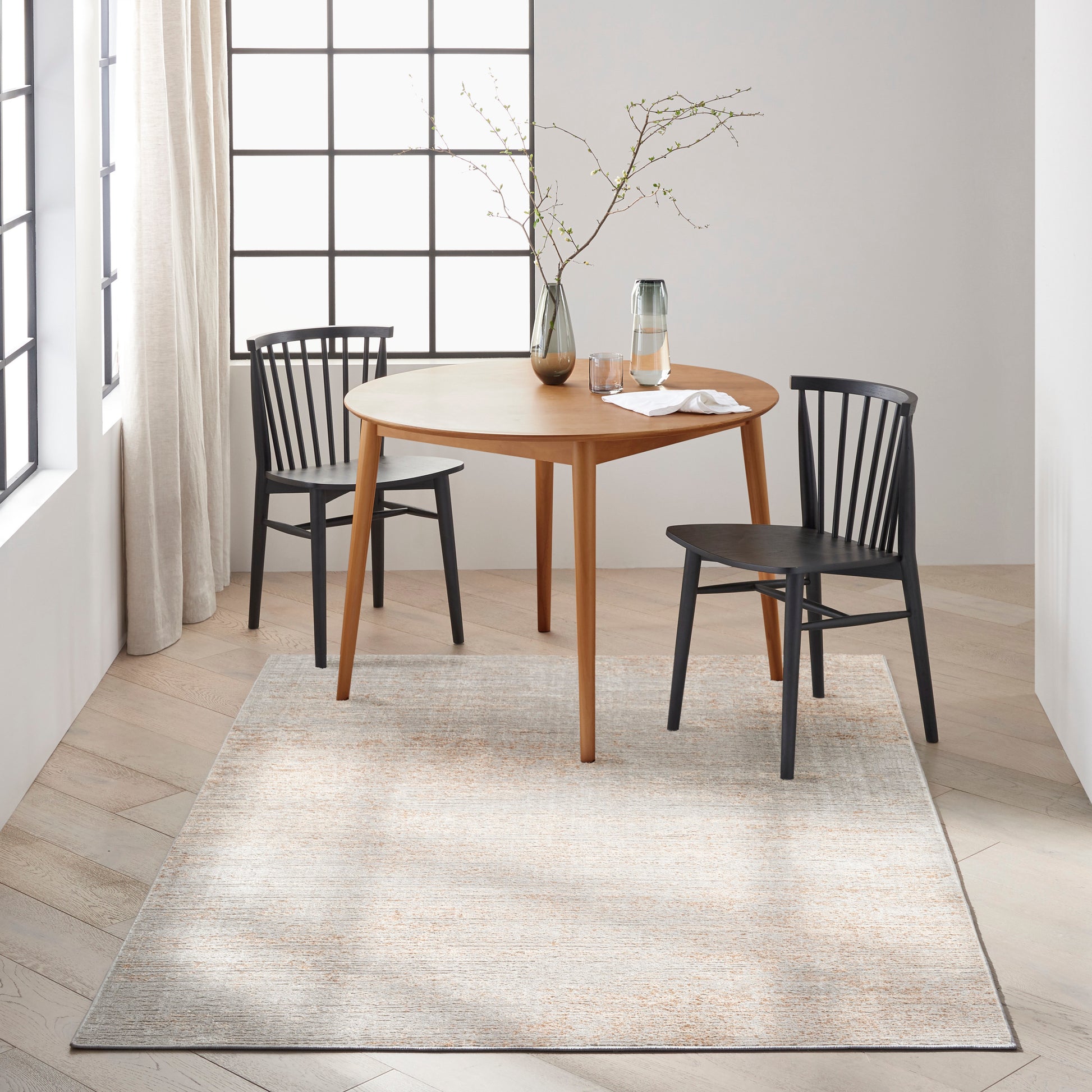 Round wooden dining table with two black chairs, glass pitcher and vase with branches on a beige rug.