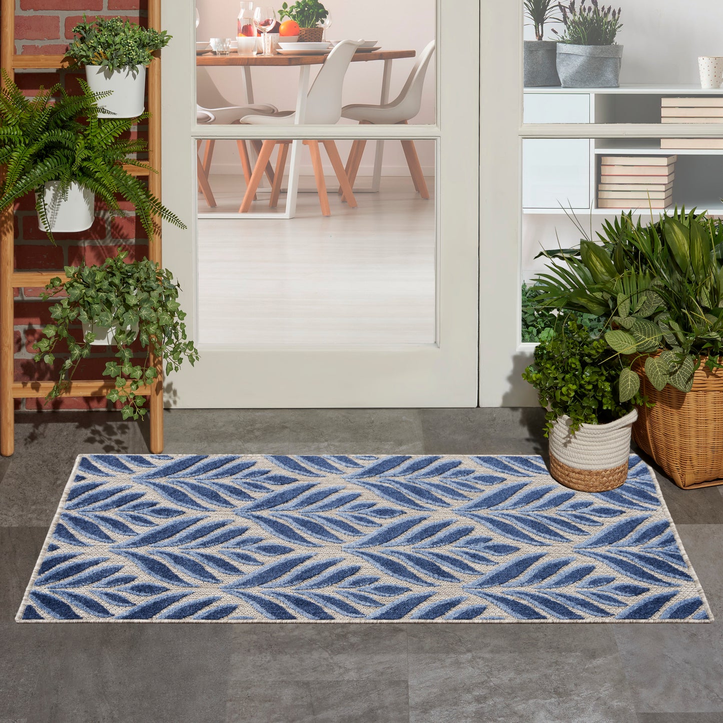 Blue and ivory leaf-patterned outdoor rug on gray tile at a glass-door entry, flanked by potted plants and a wicker basket.