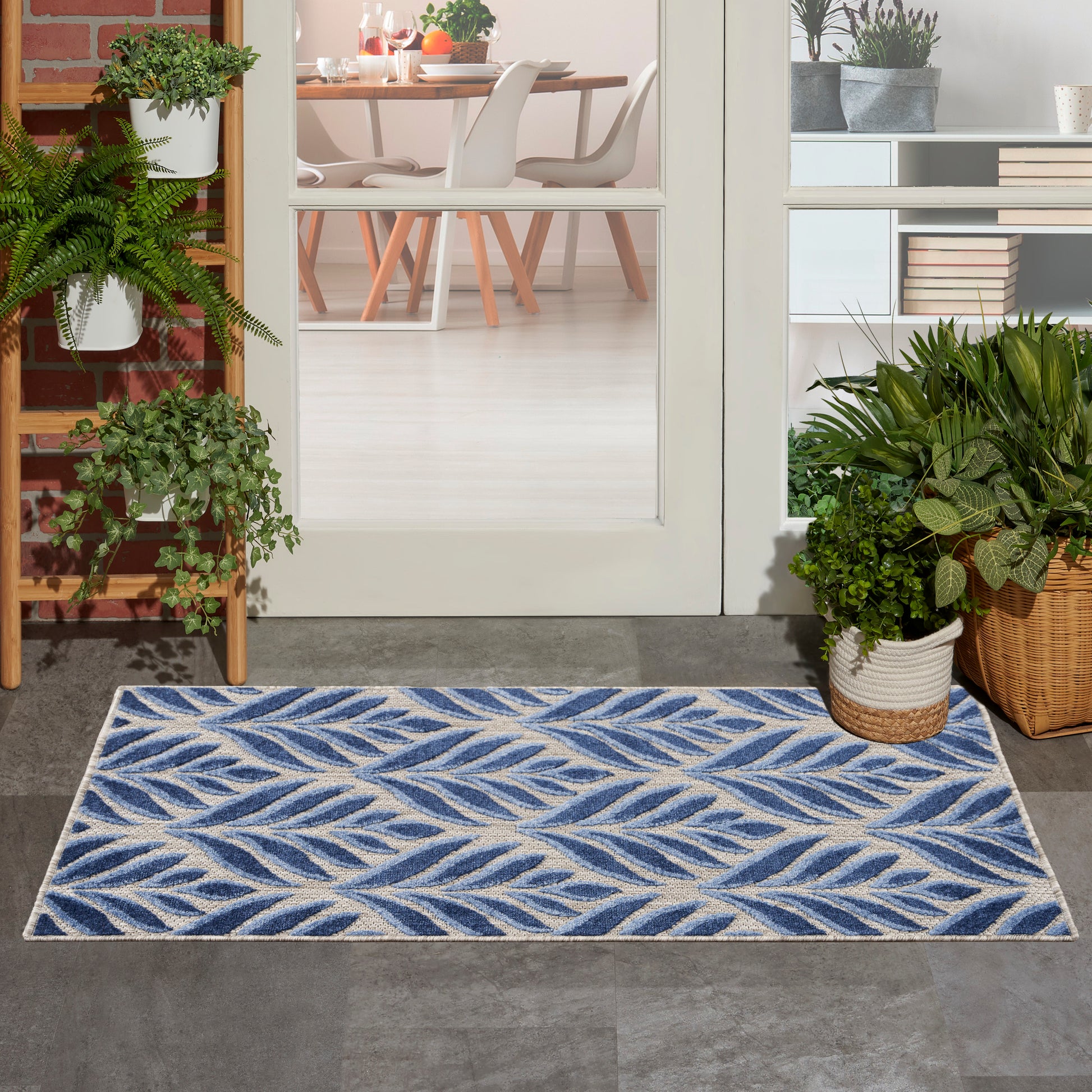 Blue and ivory leaf-patterned outdoor rug on gray tile at a glass-door entry, flanked by potted plants and a wicker basket.