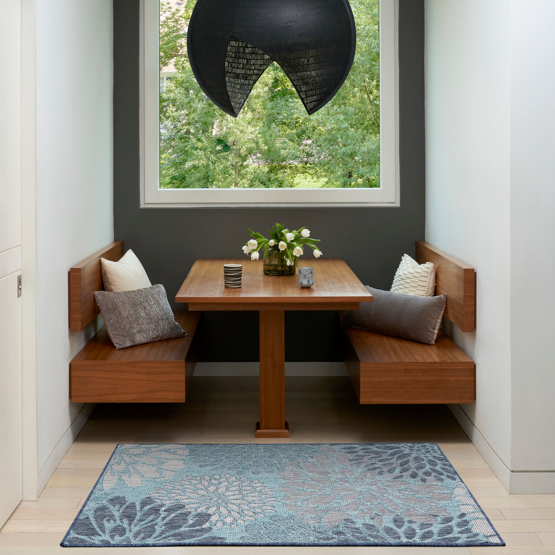 Wooden corner dining nook with L-shaped benches, square pedestal table, blue floral rug, cushions, vase of white flowers, black dome pendant.