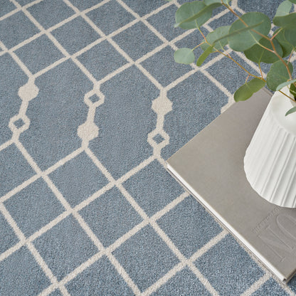 Blue-gray rug with a white geometric lattice pattern; potted plant in a white vase sits on a book beside.