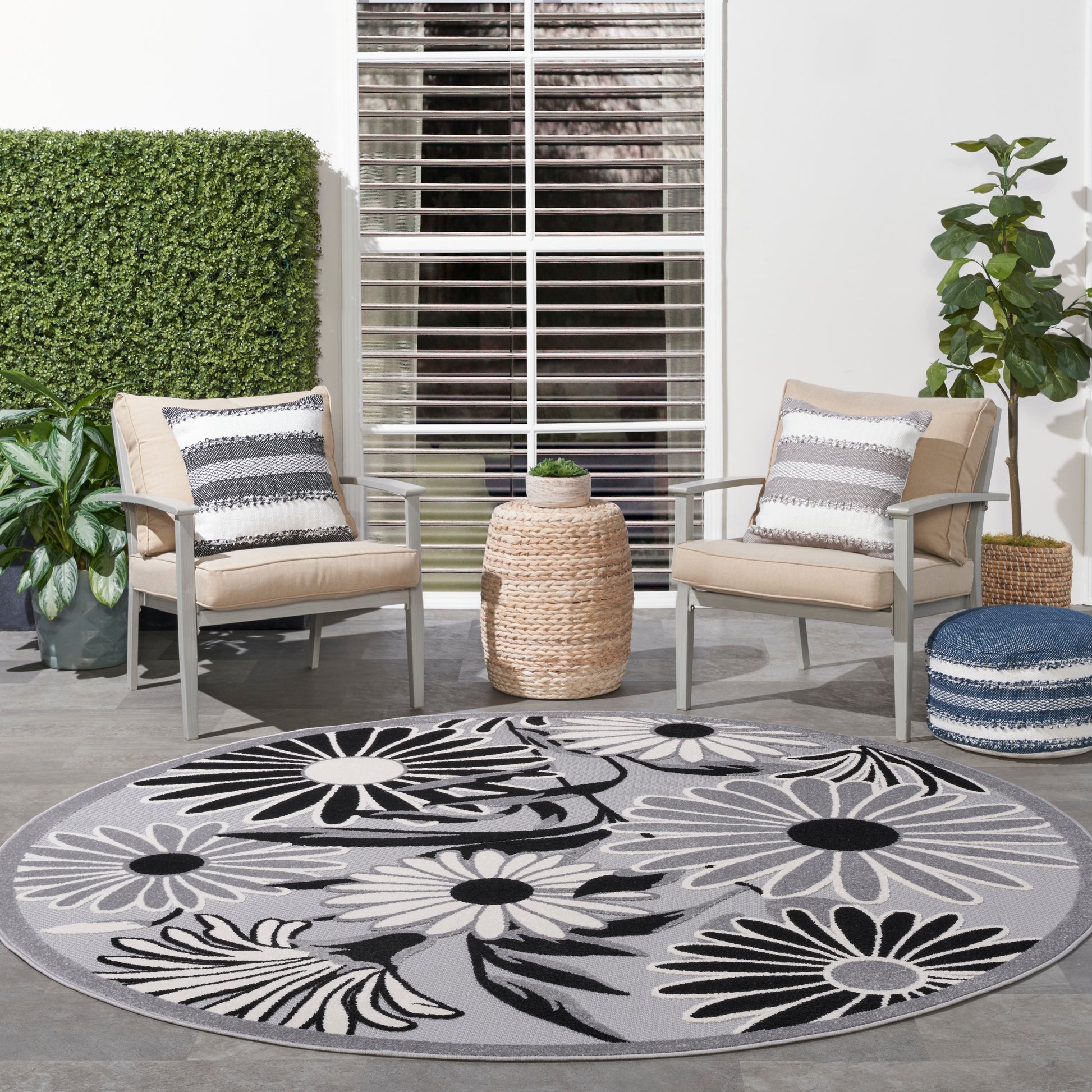 Outdoor patio with two beige cushioned chairs, round gray floral rug in black and white, woven side table, potted plants.