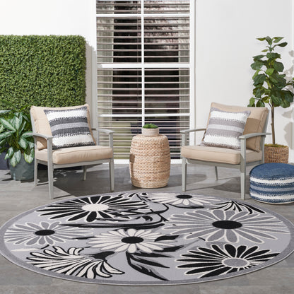 Outdoor patio with two beige cushioned chairs, round gray floral rug in black and white, woven side table, potted plants.