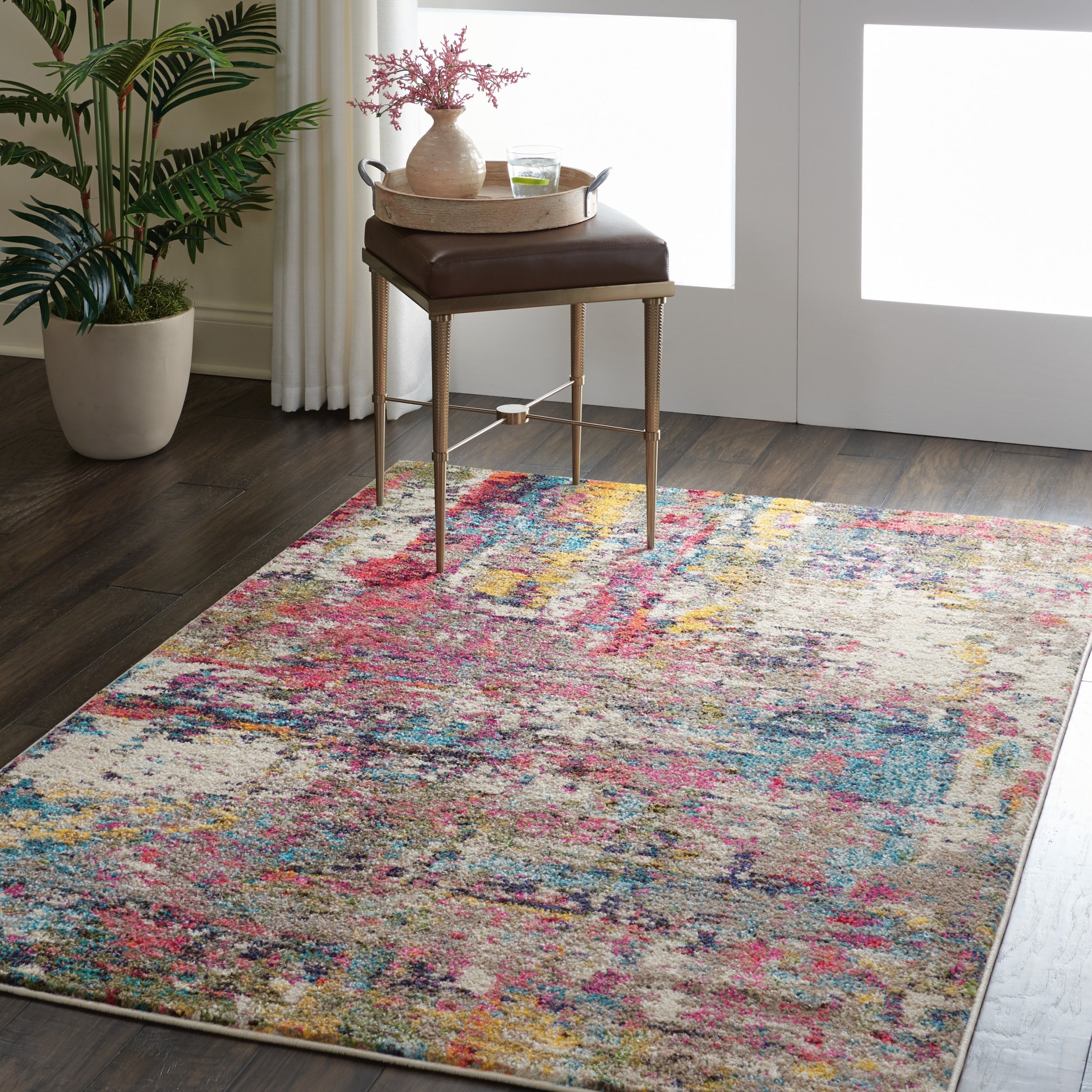 Multicolor abstract rug on dark wood floor with brown leather ottoman on brass legs, tray with vase, and green potted plant nearby.