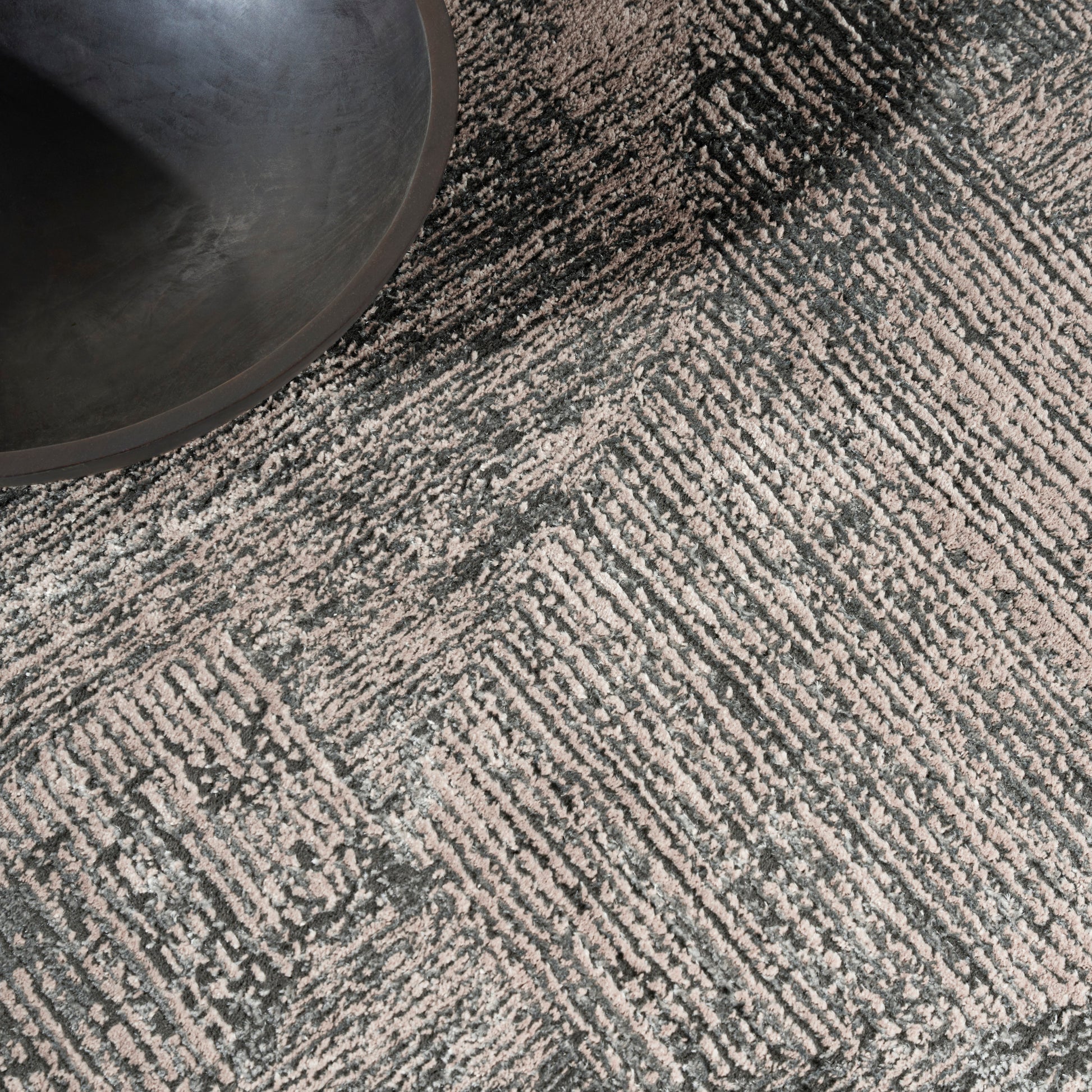 Gray textured area rug with a linear geometric pattern; a black round bowl sits on its top-left corner.