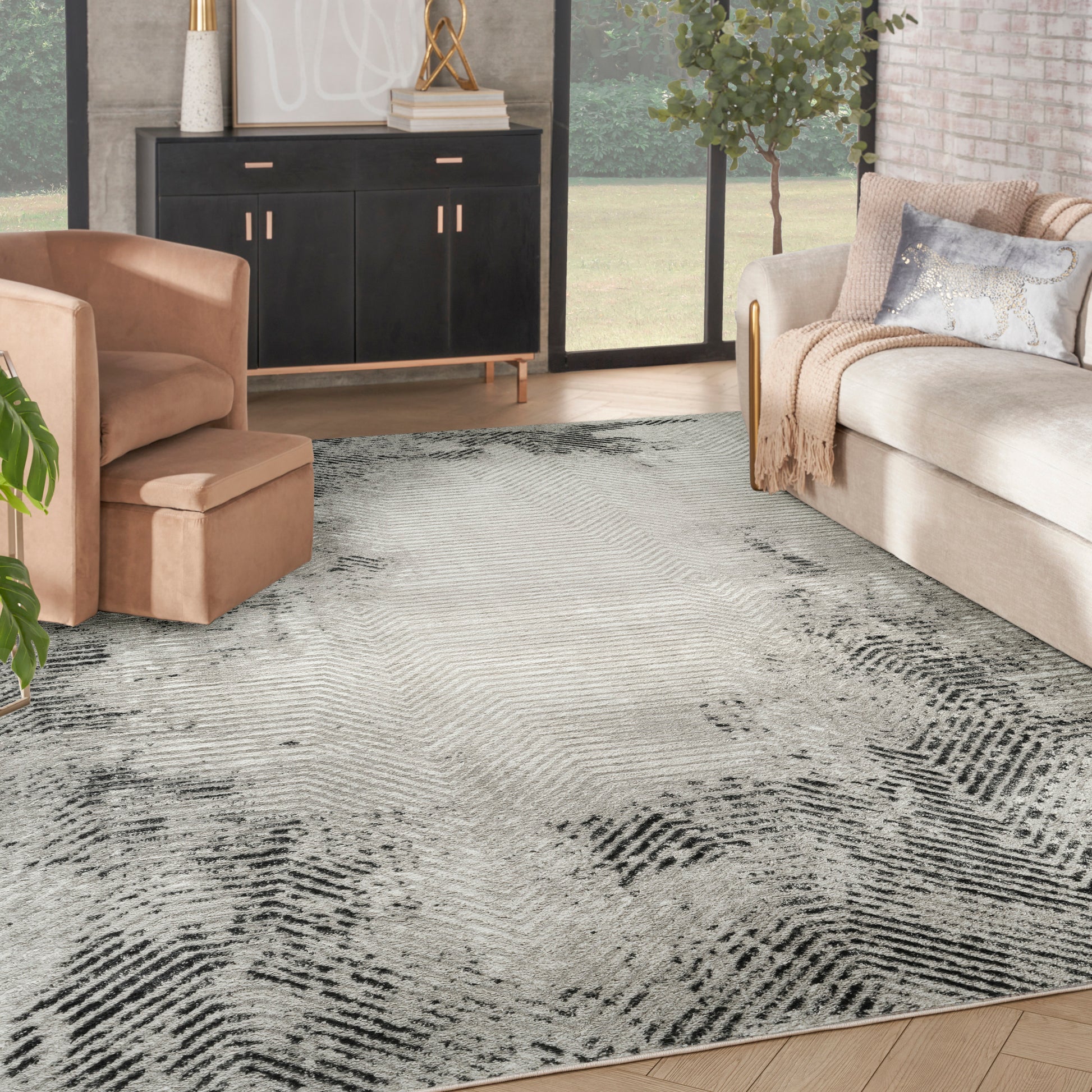 Beige sofa and tan chair with a gray patterned rug, black sideboard with gold hardware, indoor plant and large windows.