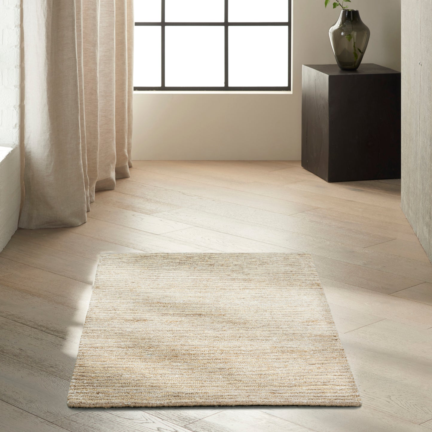 Beige woven rug on light wood floor in bright room with large grid window, cream curtains, and black pedestal vase.
