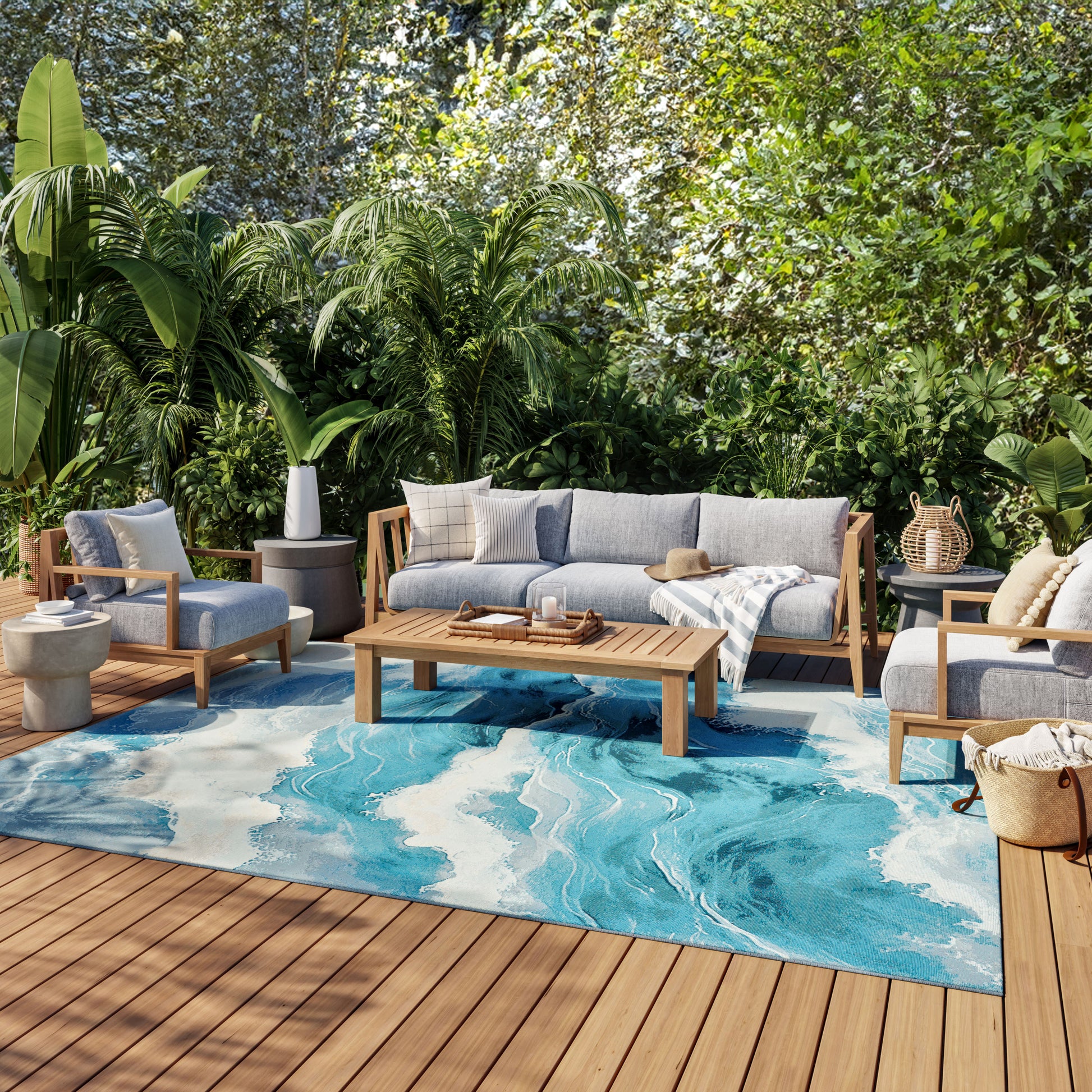 Outdoor sofa set with gray cushions on teak frames, rectangular coffee table, and a blue marble-pattern rug amid tropical plants.