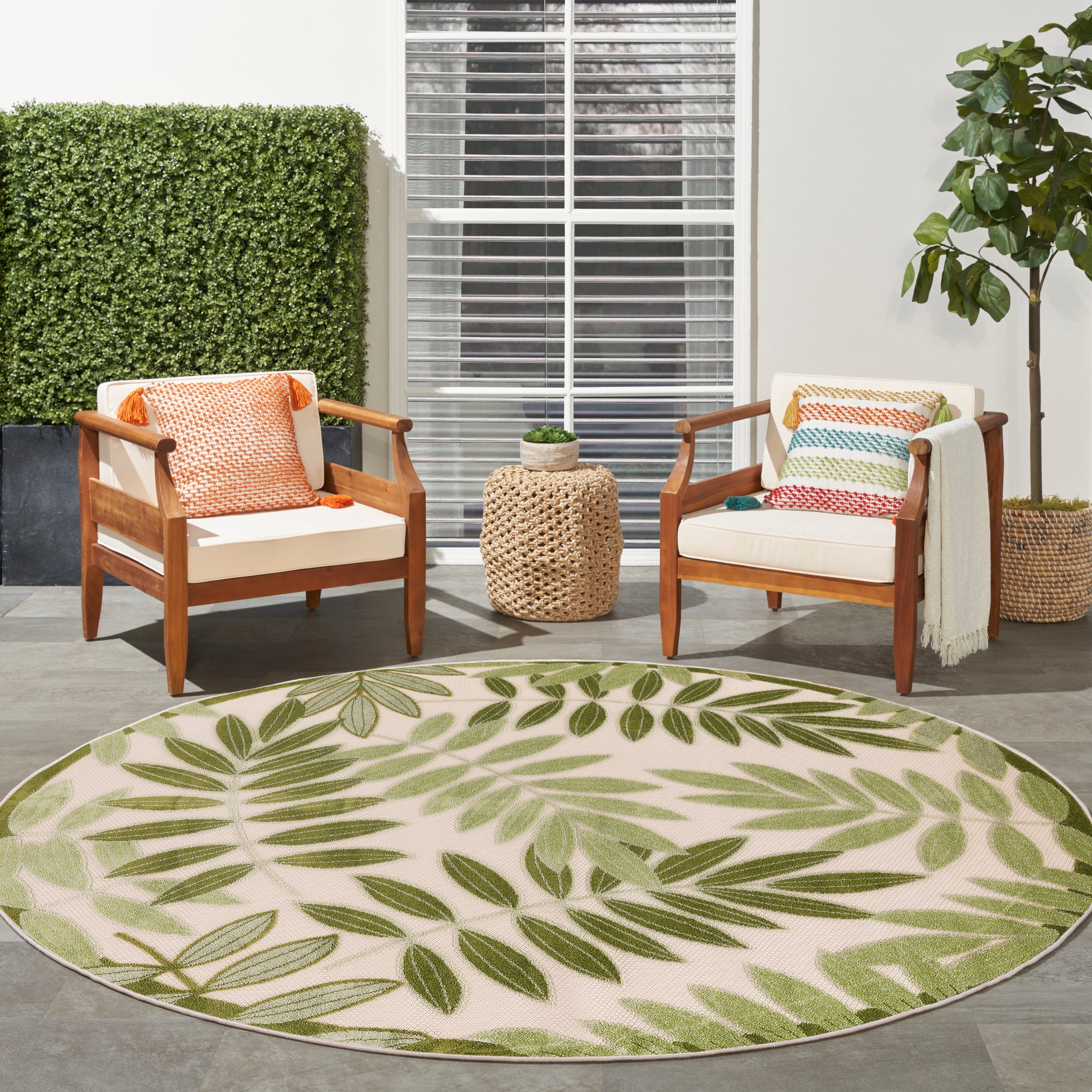 Round beige outdoor rug with green leaf pattern; wooden chairs with white cushions and colorful pillows, woven side table.