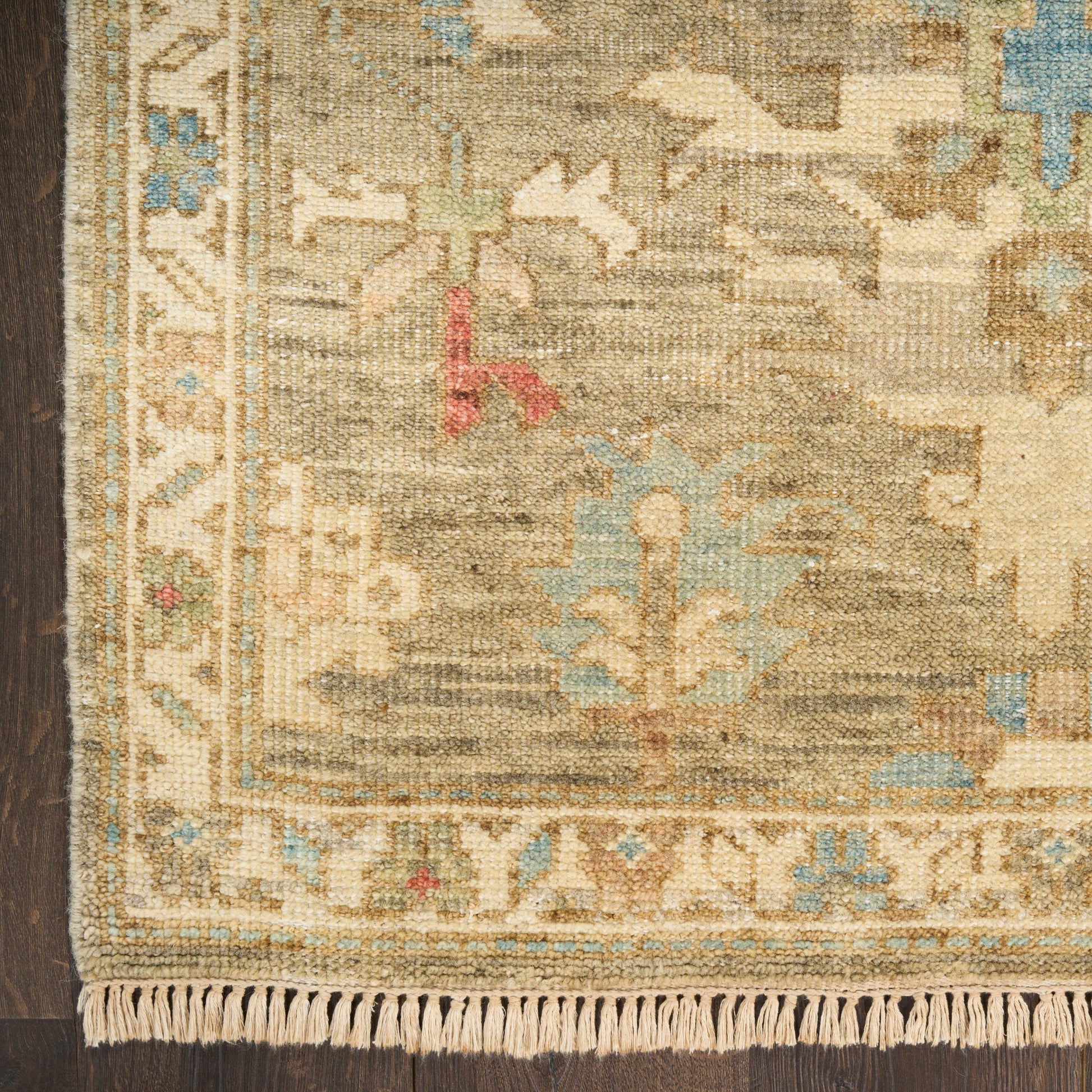 Close-up of a beige, Persian-style rug with blue accents, ornate border and fringed edge on a dark wood floor.
