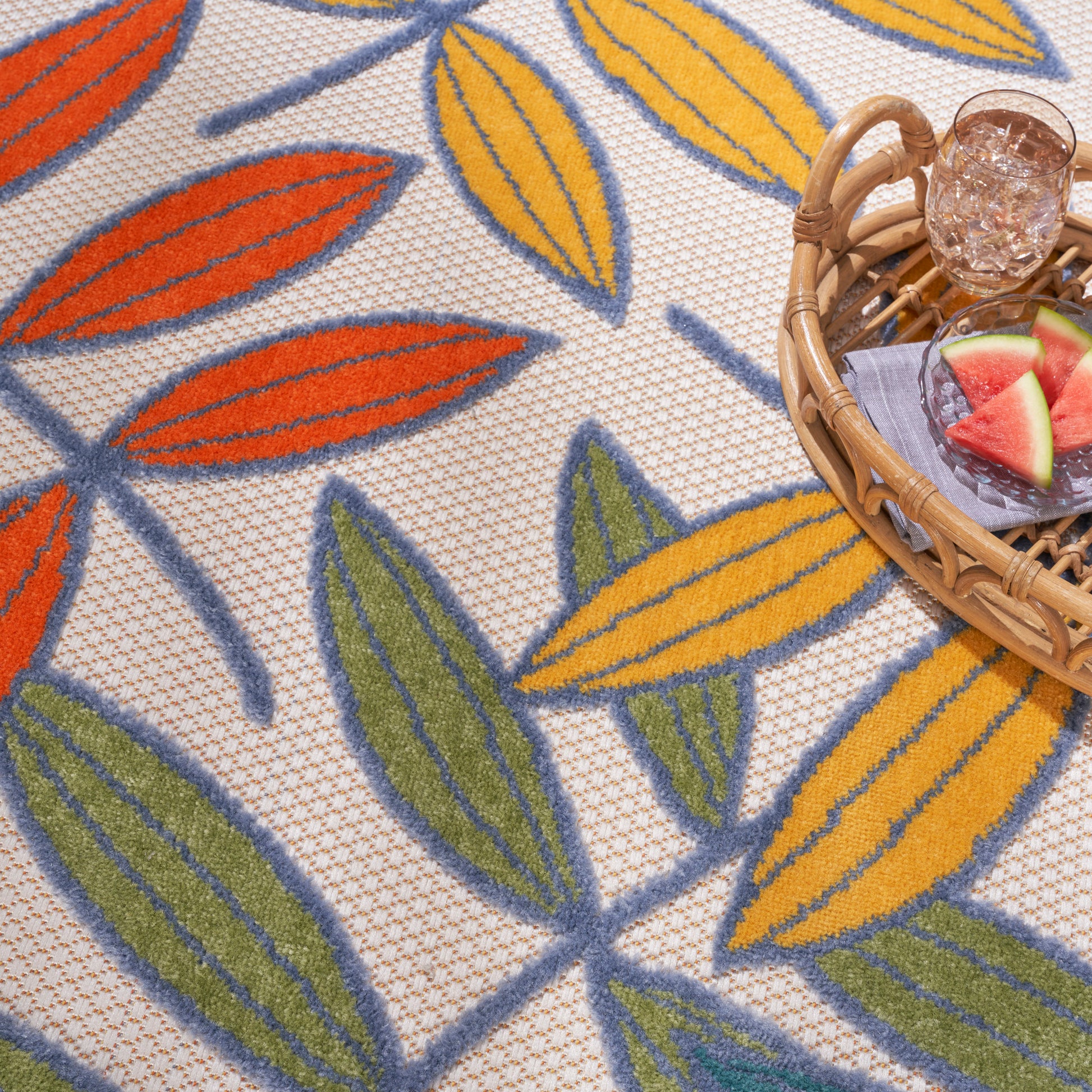 Colorful leaf-patterned woven rug in orange, yellow and green on beige; wicker basket with glass and watermelon slices.