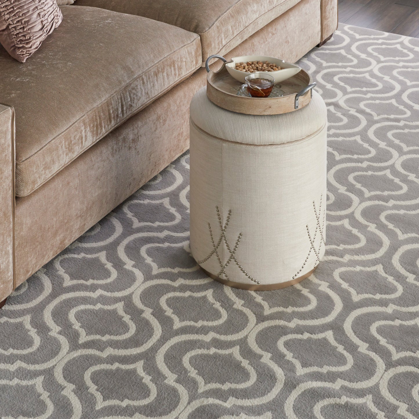 Gray trellis rug under a beige tufted sofa; beige cylindrical fabric pouf with a wooden base & metal tray sits beside the sofa.