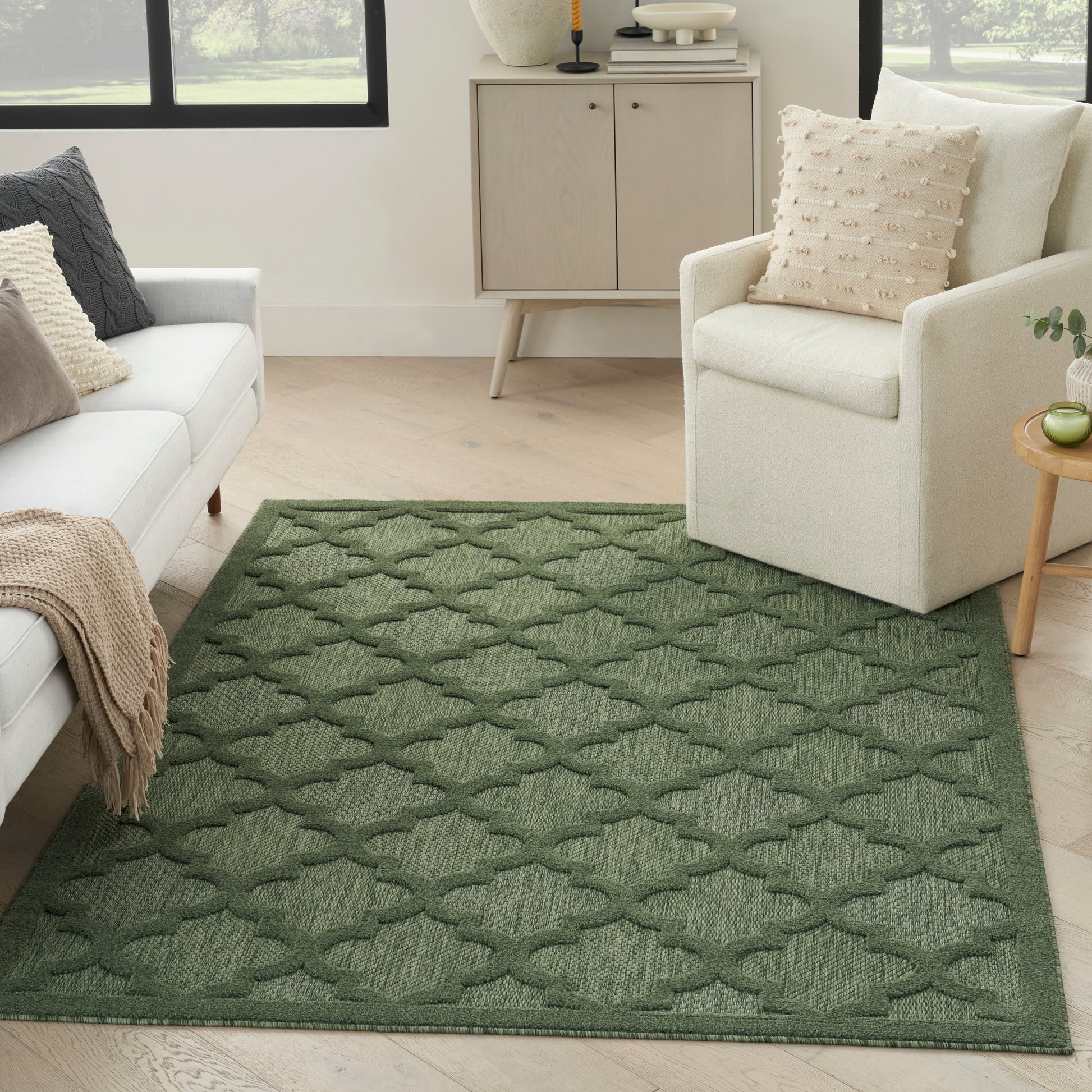Rectangular green woven rug with a repeating lattice pattern in a bright living room with a white sofa and cream armchair.