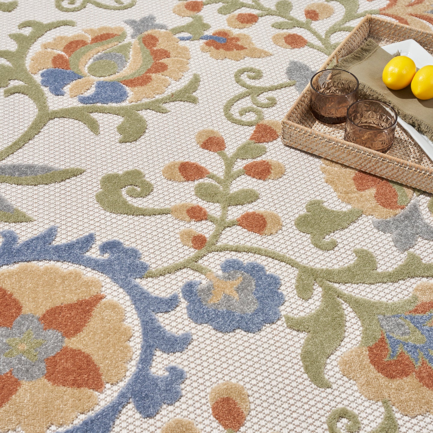 Outdoor colorful floral rug with beige background, green vines, orange & blue flowers; wicker tray with two amber glasses and lemons.