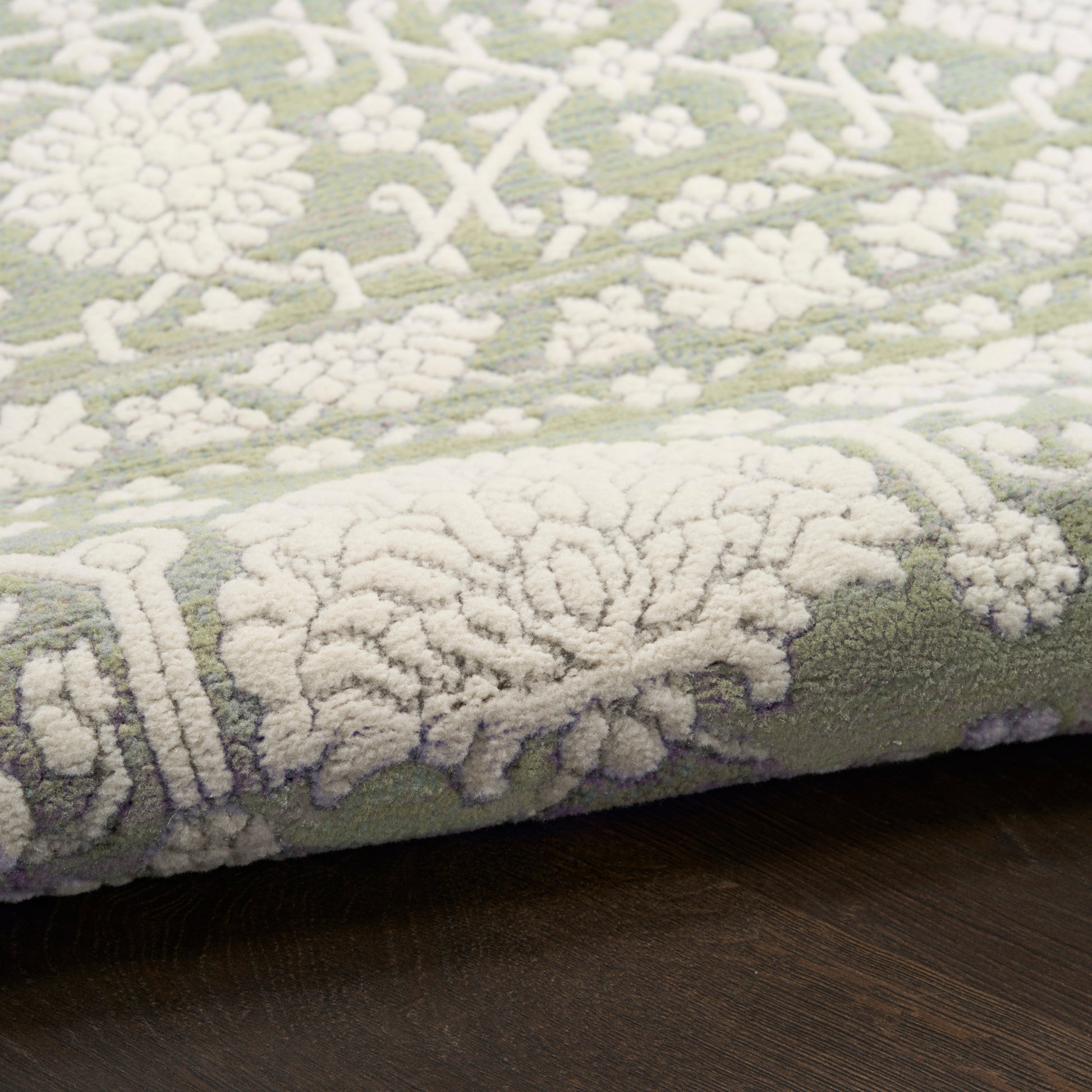Olive green rug with white raised floral damask pattern, soft plush pile on dark wooden floor.