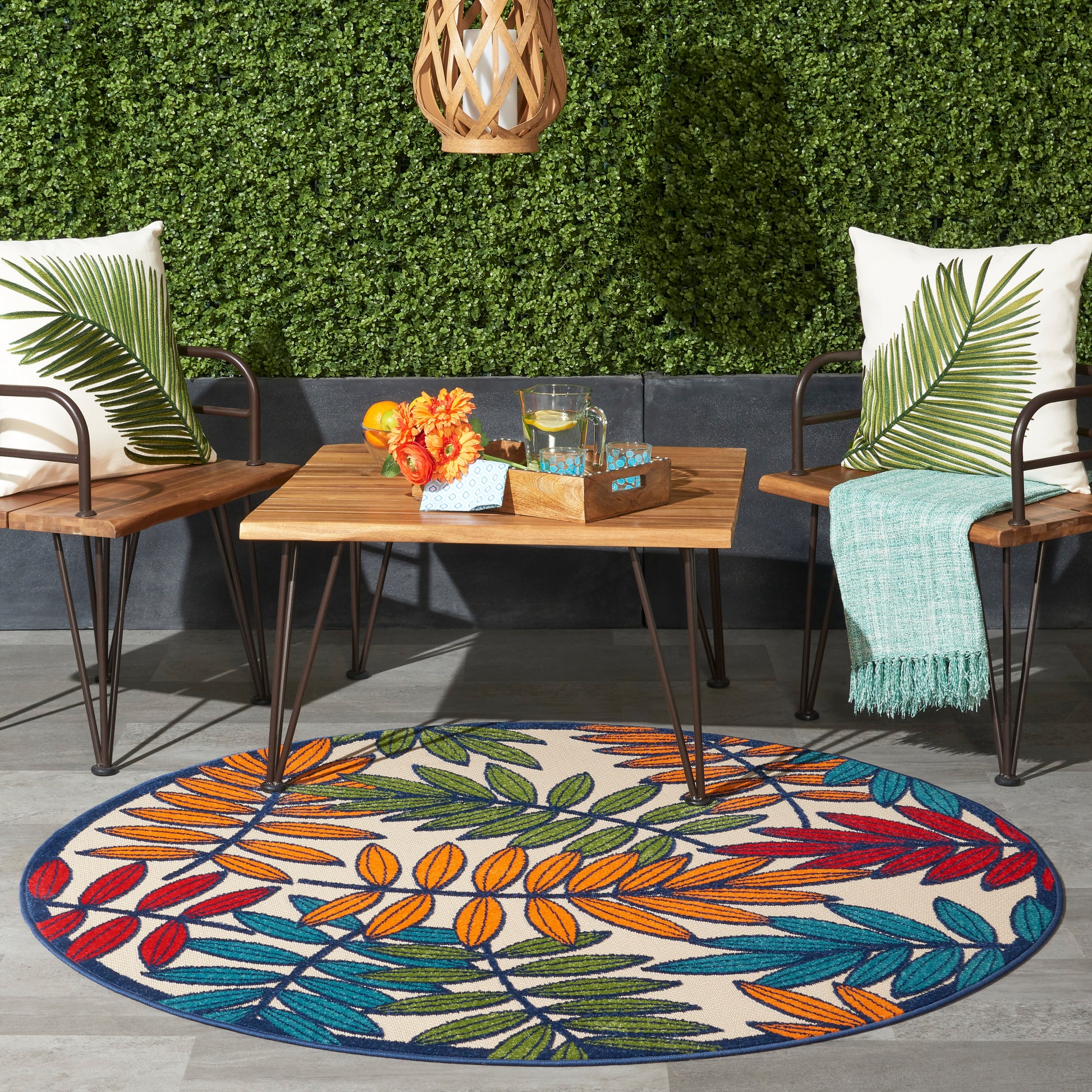 Round outdoor rug with bright multicolor leaf pattern in orange, blue, green under a wooden coffee table and wicker chairs.