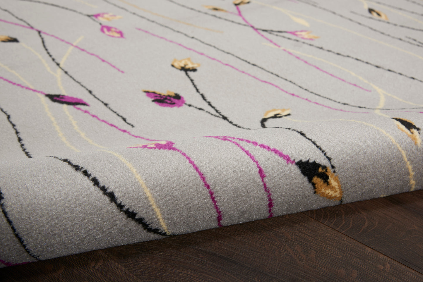 Grey area rug with abstract pink, yellow & black lines and small floral accents, folded edge on wood floor.