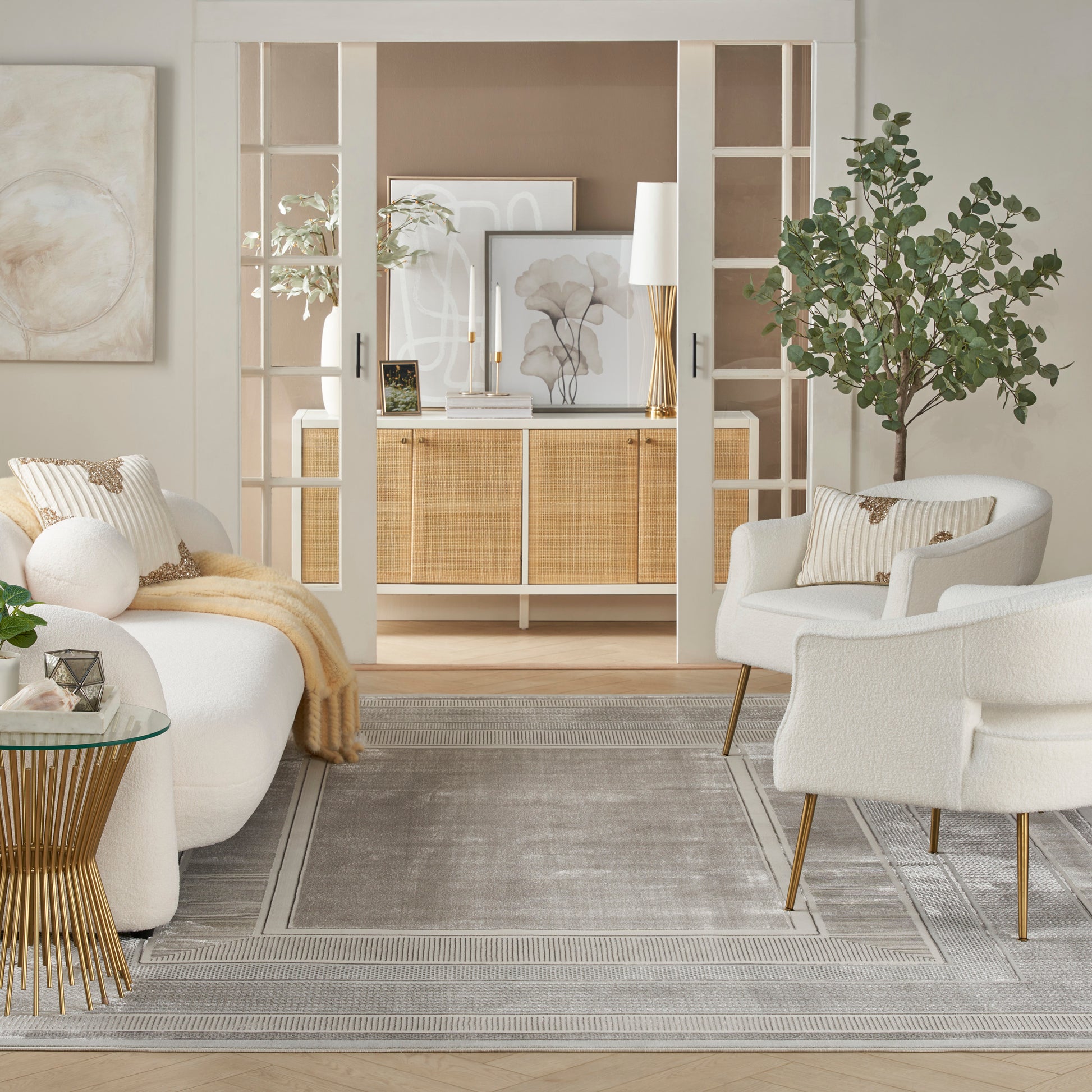 Neutral living room with white boucle chairs, glass-topped gold table, gray rug, beige throw, and woven rattan cabinet.