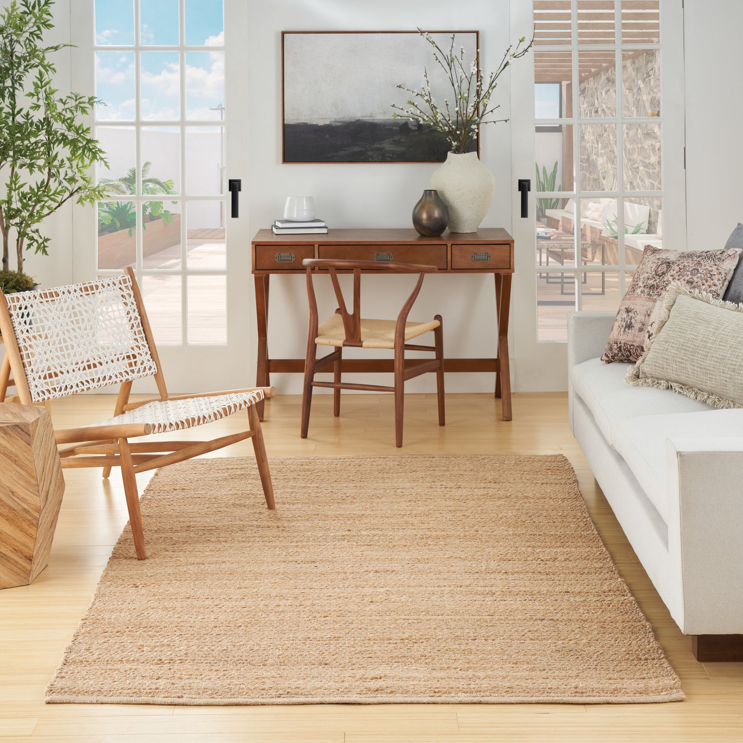 Bright living room with natural jute rug, mid-century wooden desk & chair, woven lounge chair, white sofa, sunlit windows.