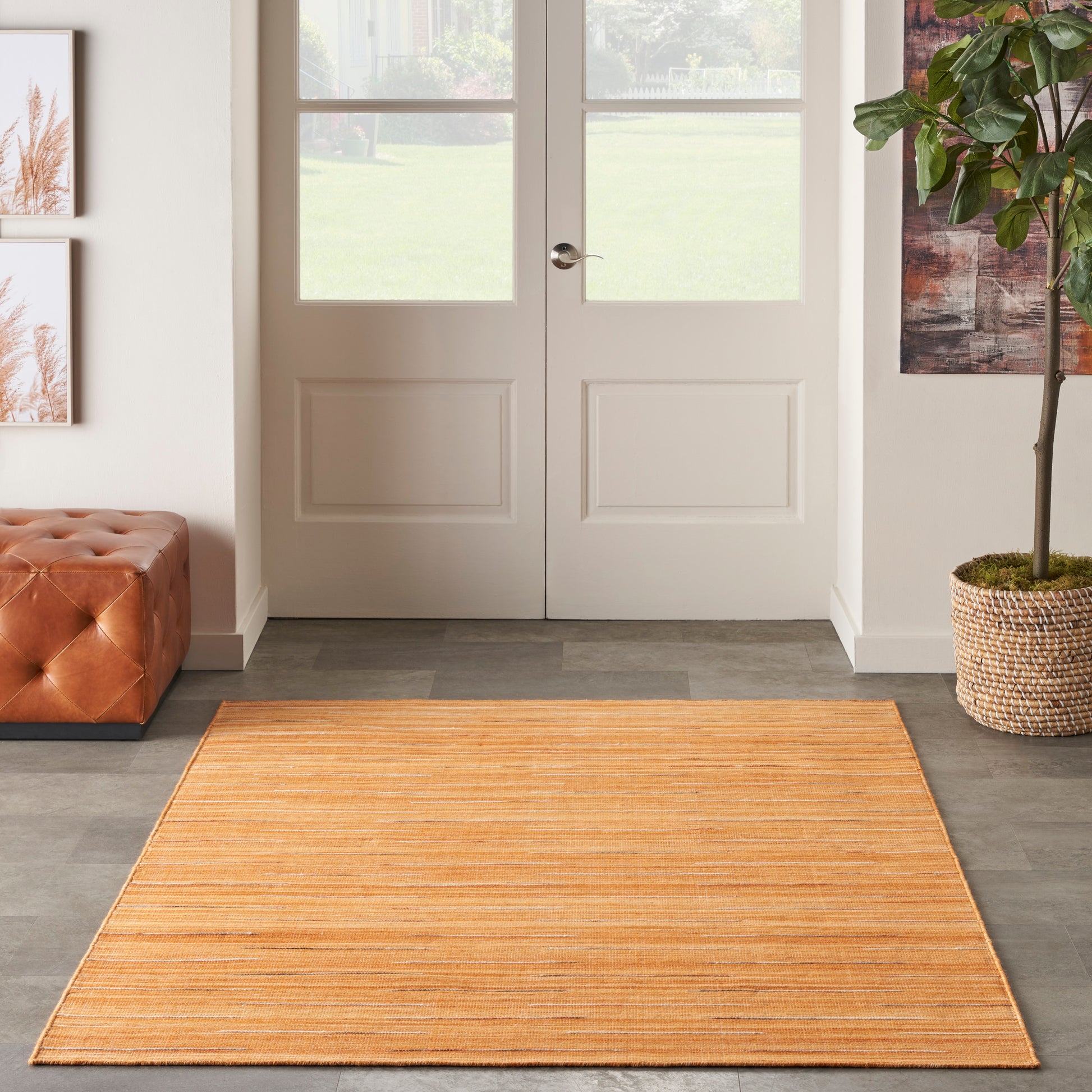 Rectangular natural fiber orange rug in a modern entryway with white paneled doors, a tufted brown ottoman, and a potted plant.