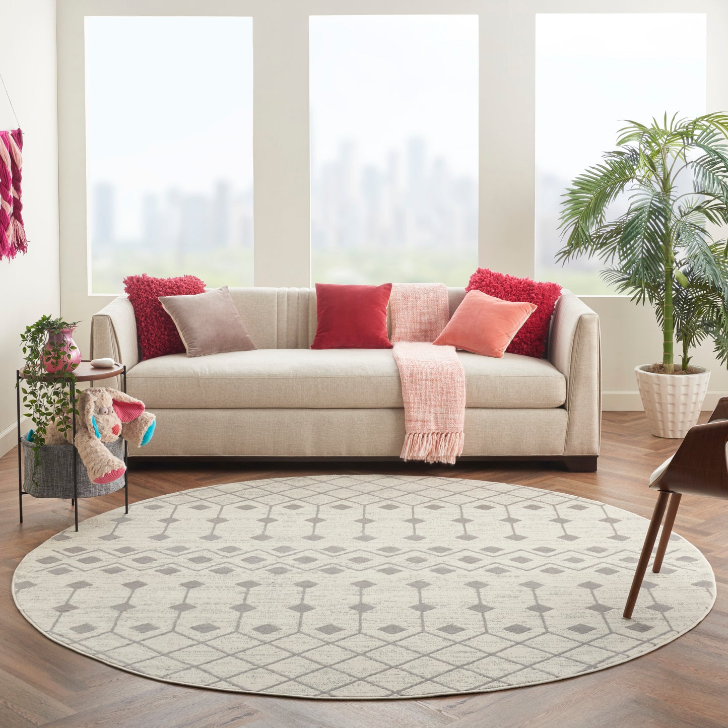 Round grey rug in a beige living room with a beige fabric sofa, red & pink cushions, pink throw, plants, and city-view windows.