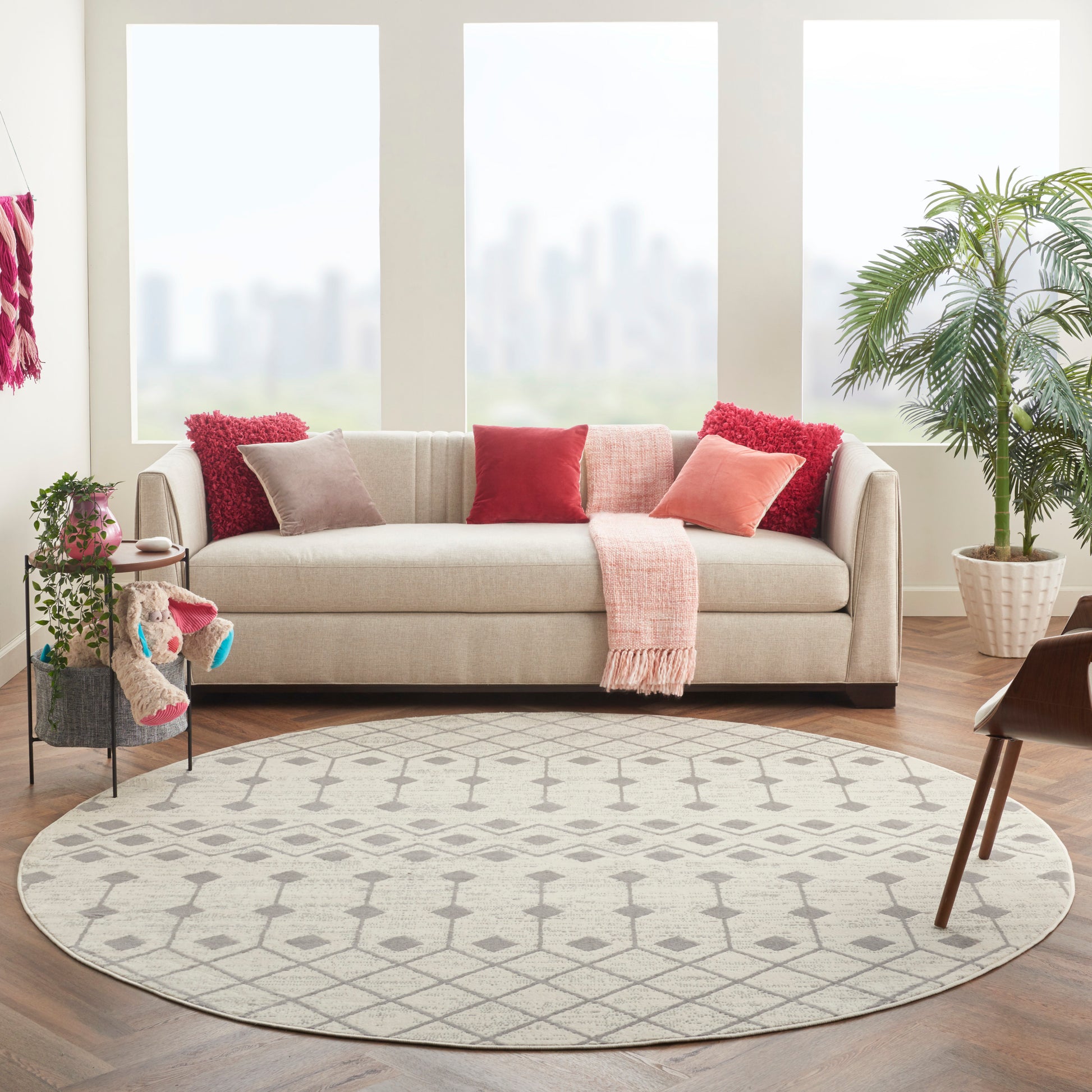 Round grey rug in a beige living room with a beige fabric sofa, red & pink cushions, pink throw, plants, and city-view windows.