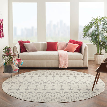 Round grey rug in a beige living room with a beige fabric sofa, red & pink cushions, pink throw, plants, and city-view windows.