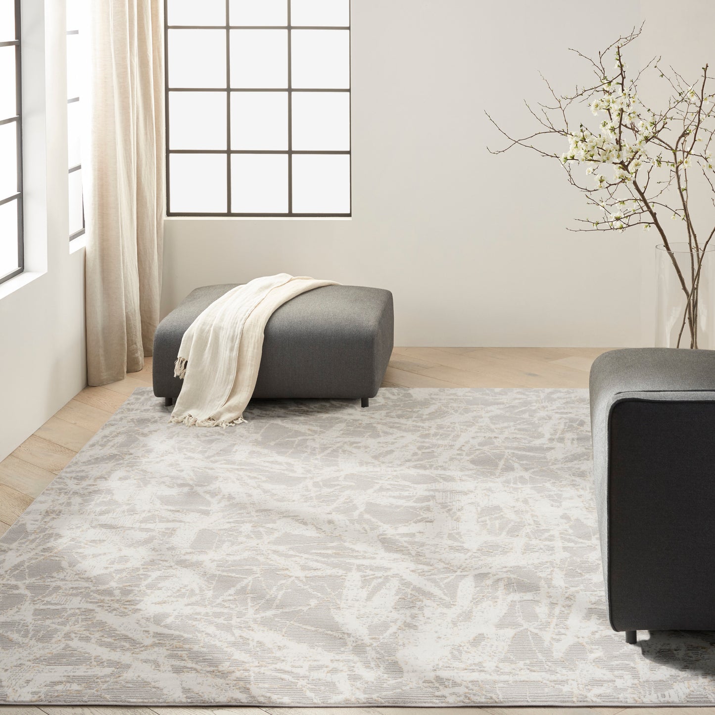 Light gray abstract-pattern area rug with two dark gray ottomans, cream throw, in a bright modern living room.
