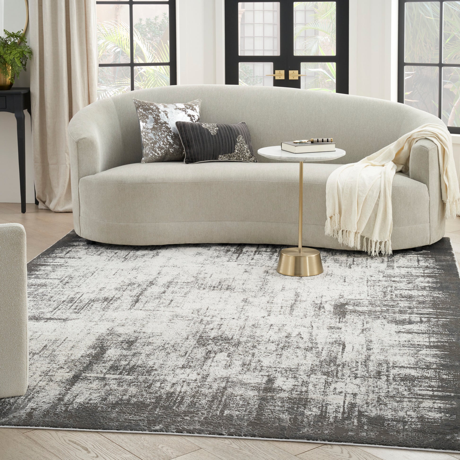 Curved beige fabric sofa with ivory throw, gold side table, metallic-pattern cushions, and a grey abstract rug.