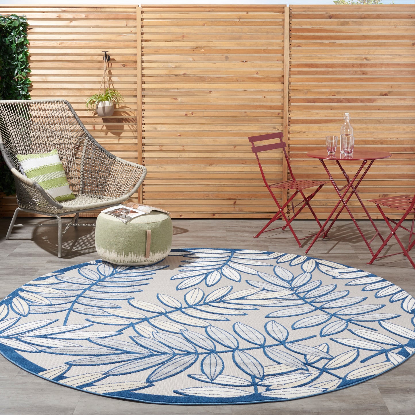 Round blue-and-white leaf-pattern rug on a wooden deck with a wicker chair, green pouf, and red bistro table and chairs.