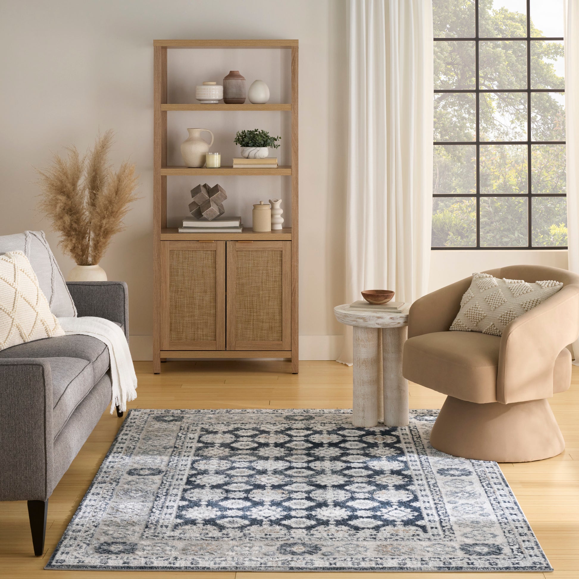 Cozy living room with a tall wood shelf, beige rounded armchair, gray sofa, blue patterned rug, pampas vase, and round table.