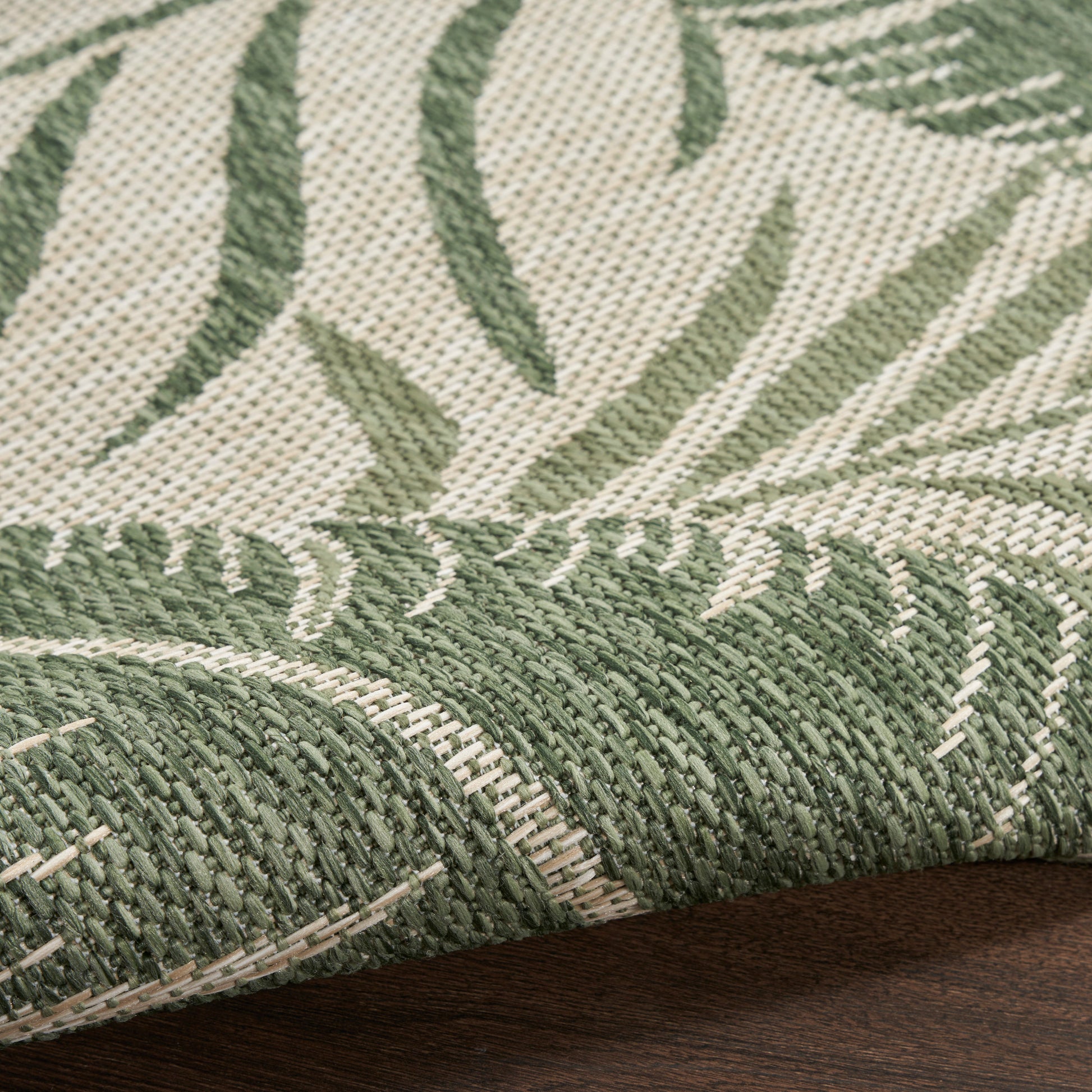Woven rug with olive-green botanical leaf pattern on beige background; textured, rolled edge exposed.