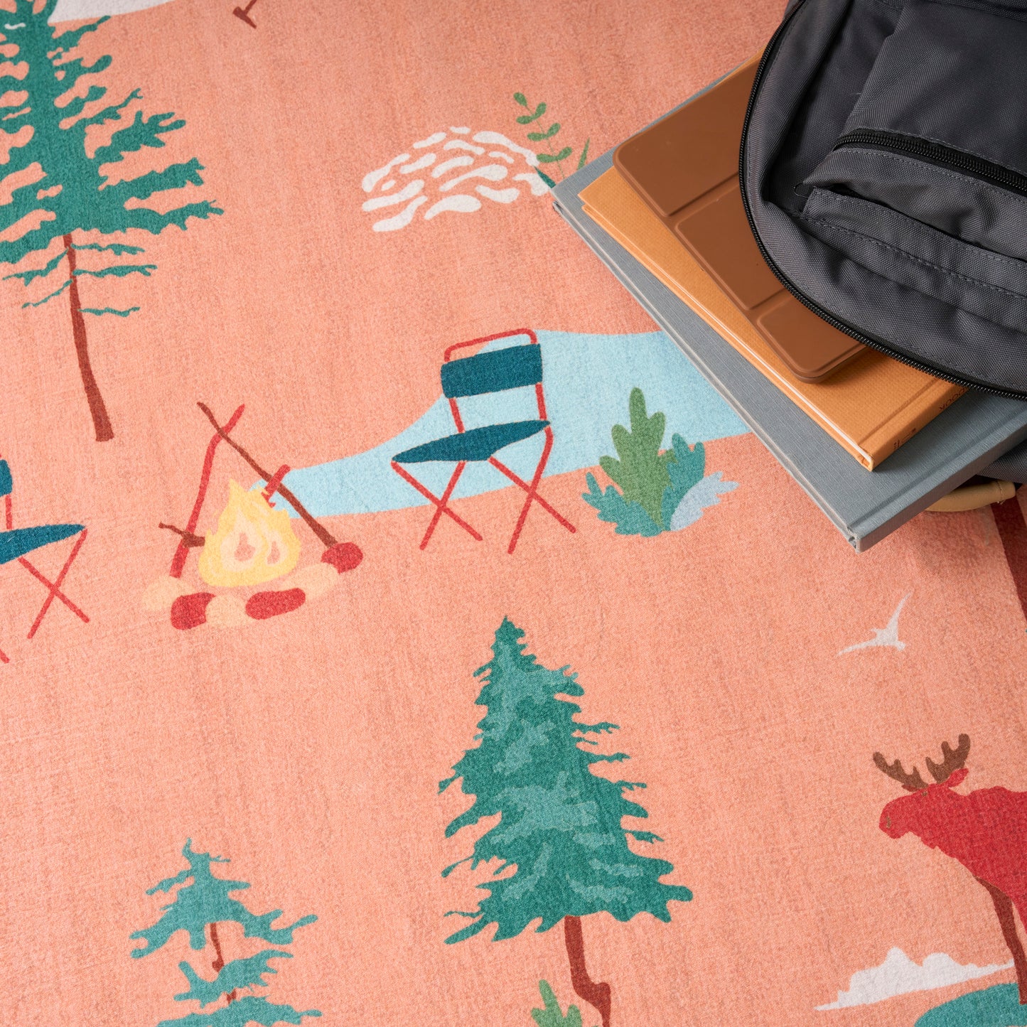 Peach camping rug with trees, campfire and blue chair print; black backpack atop brown notebooks.