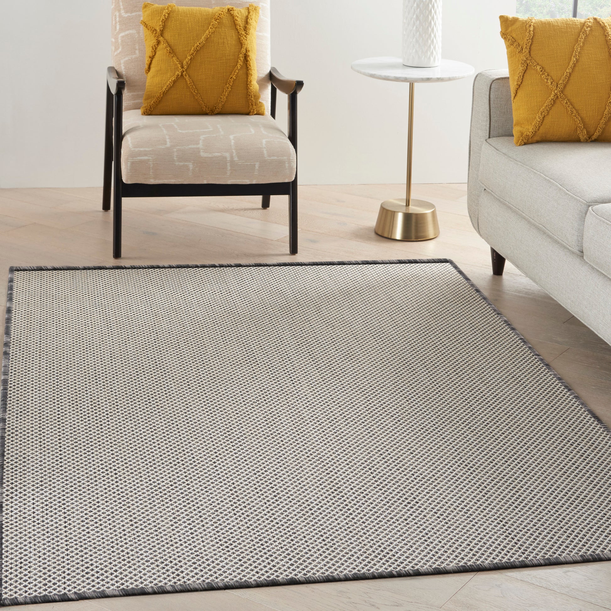 Grey woven area rug with subtle geometric pattern & dark border in a modern living room with beige armchair & mustard throw pillows & a round brass-topped side table.