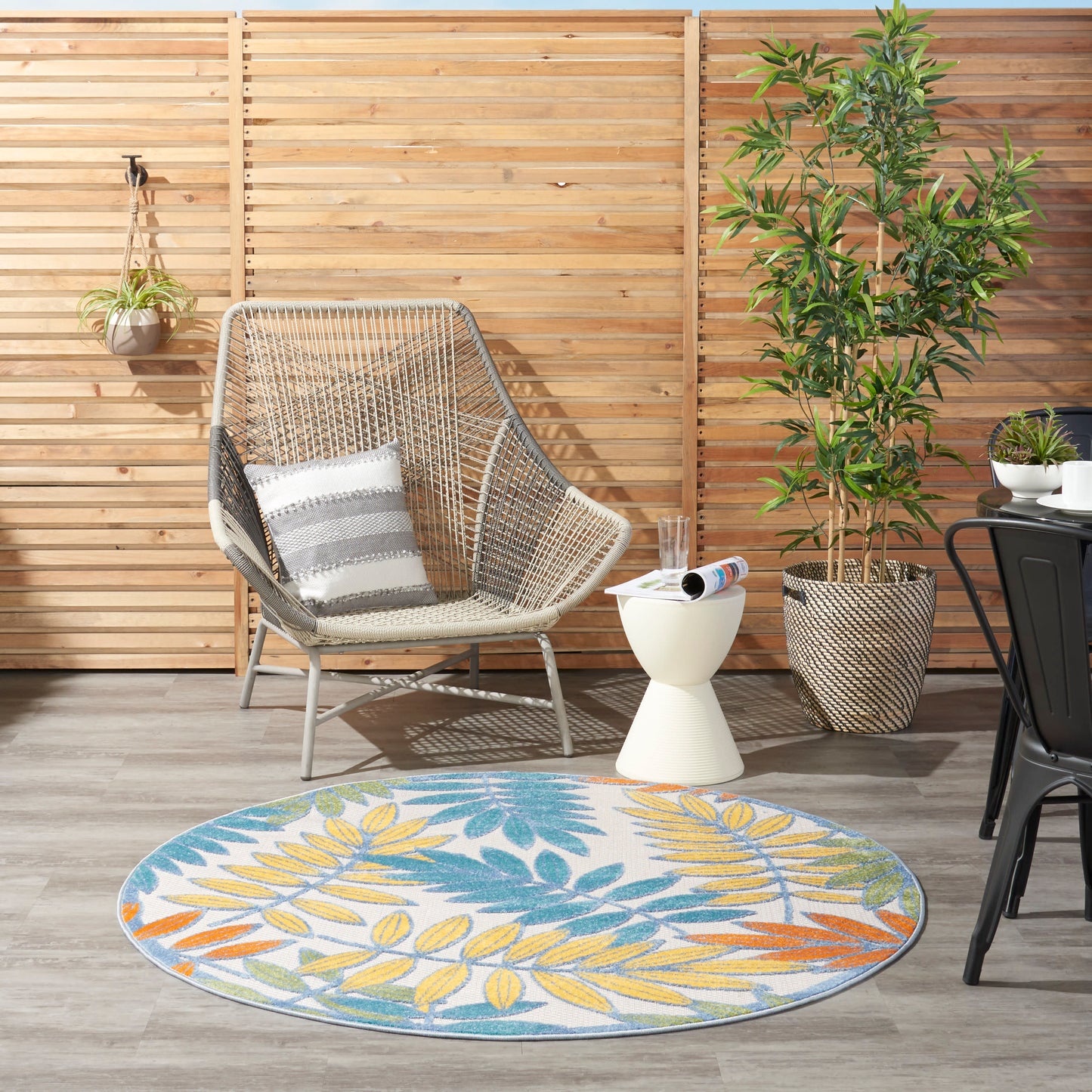 Outdoor patio with a woven chair, white cushion, round colorful leaf rug, potted plant, side table, and wooden slat fence.