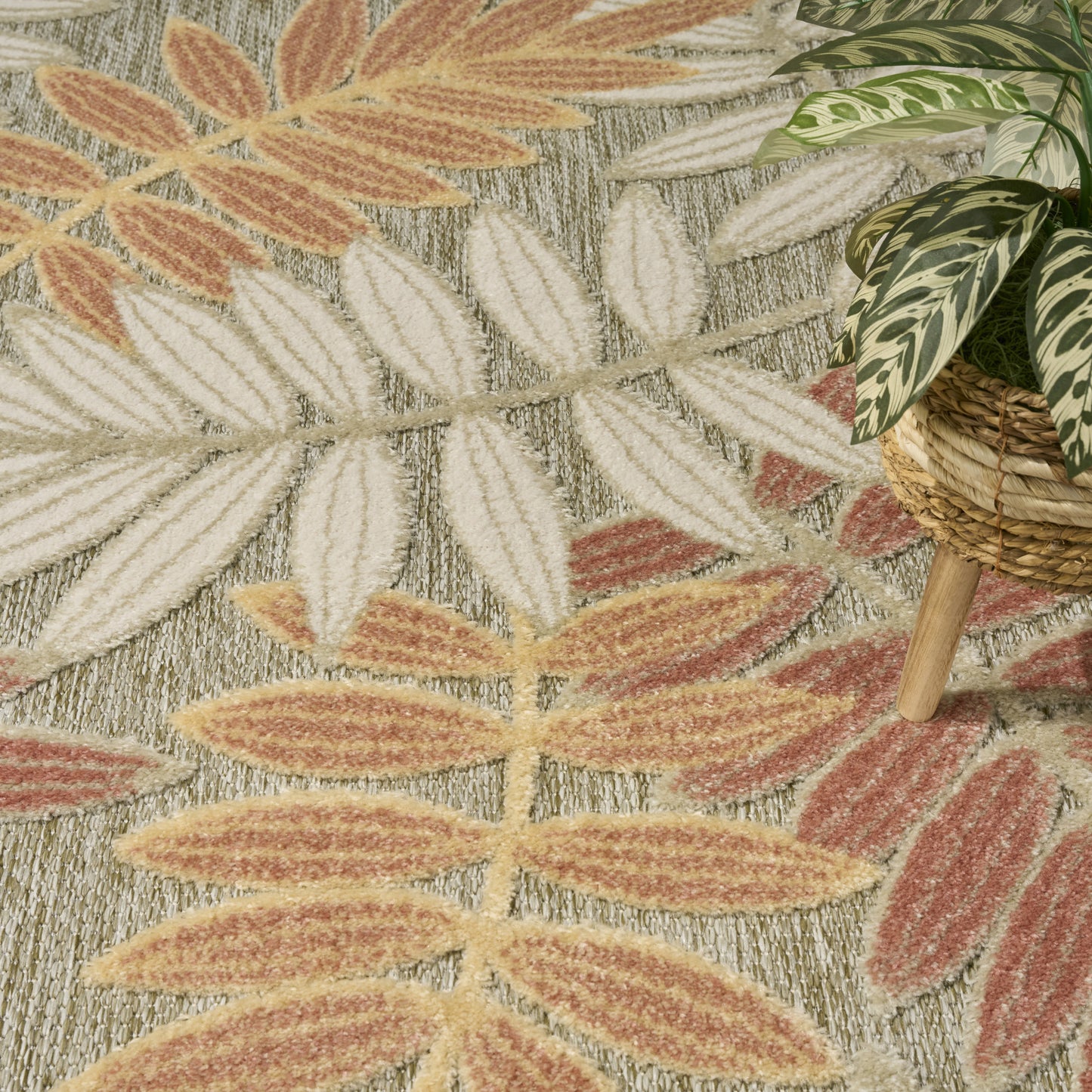 Textured area rug with multicolored leaf patterns in cream, peach, terracotta, sage; woven fiber look, plant in basket nearby.