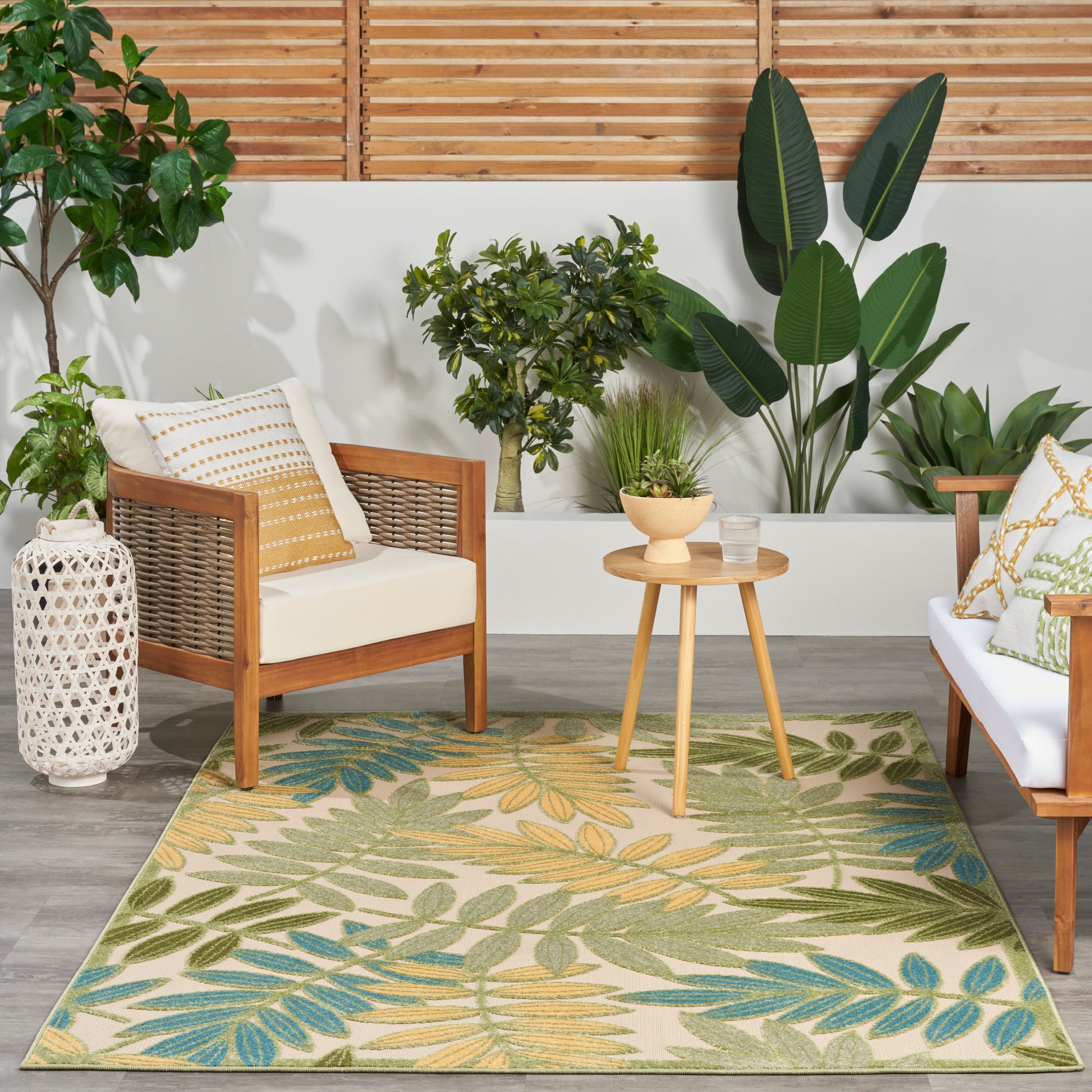Boho outdoor lounge with wicker chairs, cream cushions, round wooden side table, and tropical leaf rug amid lush plants.