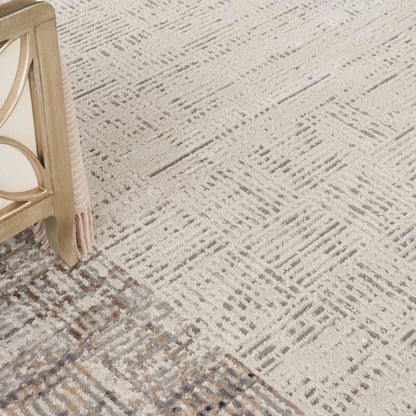 Beige-taupe geometric area rug with gray accents, beside pale wood furniture leg and pink fringe detail.