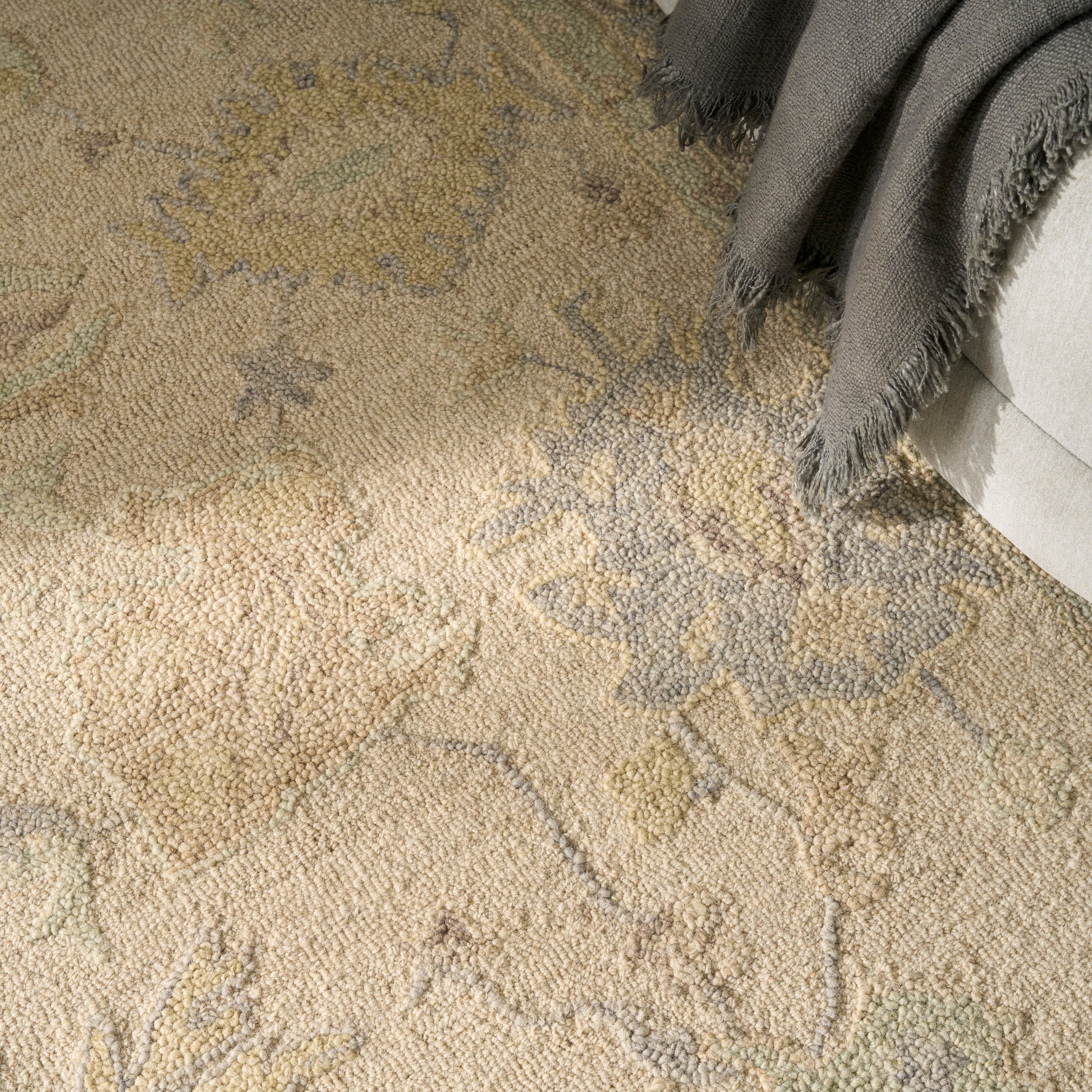 Beige textured area rug with blue-gray floral motif; soft loop pile, gray throw blanket at sofa edge.