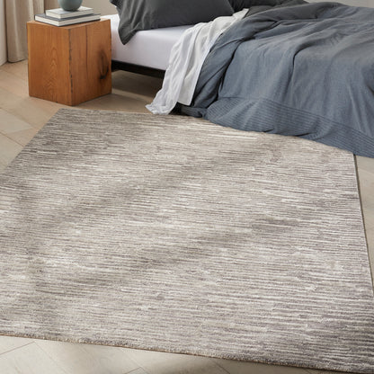 Rectangular beige-gray textured area rug on light wood floor beside bed with gray bedding and a wooden nightstand.