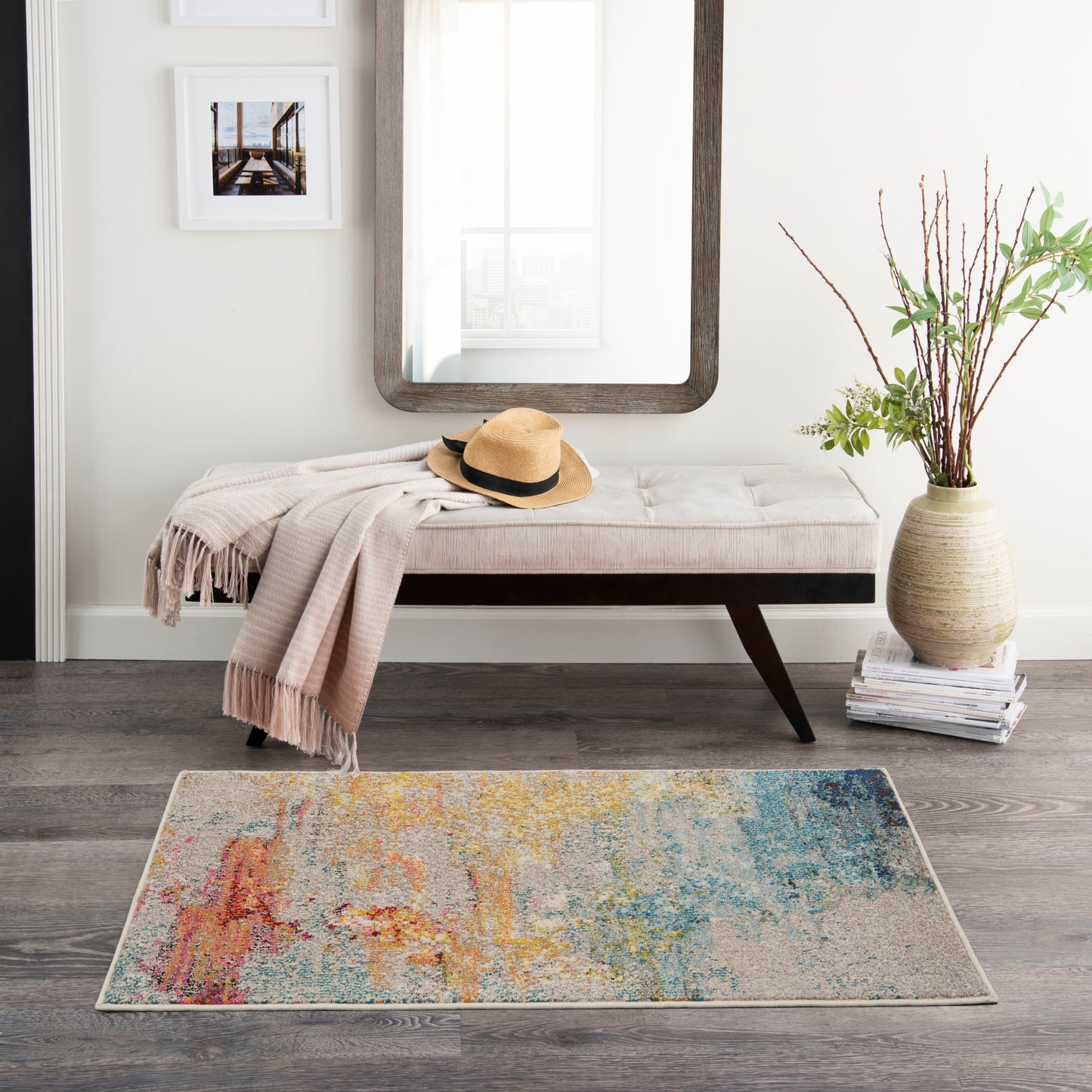 Modern entryway with beige tufted bench, straw hat, blush throw, rounded mirror, colorful abstract rug, vase with branches.