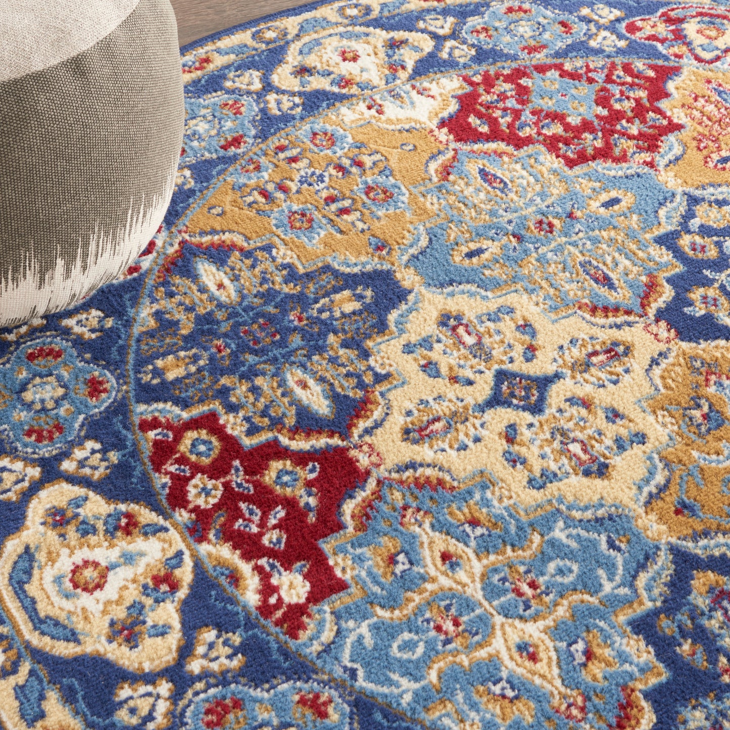 Close-up of a Persian-style area rug in blue, ivory, red & gold with floral medallions; light gray ottoman visible.