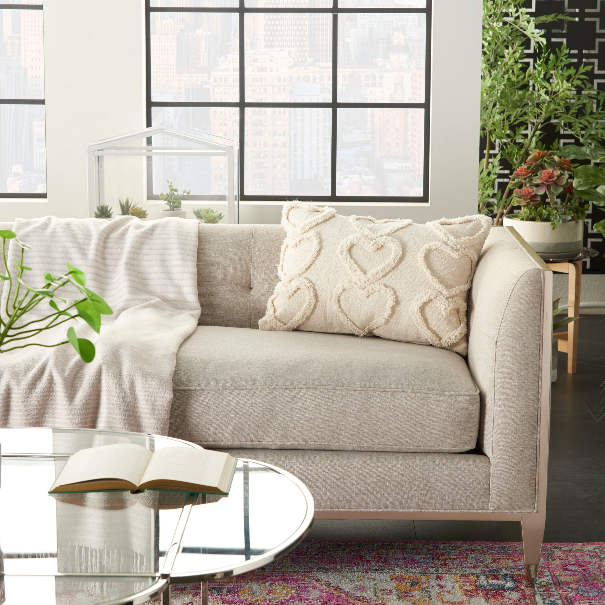 Beige fabric sofa with heart-patterned cream cushions, glass round coffee table, linen throw, bright urban living room.