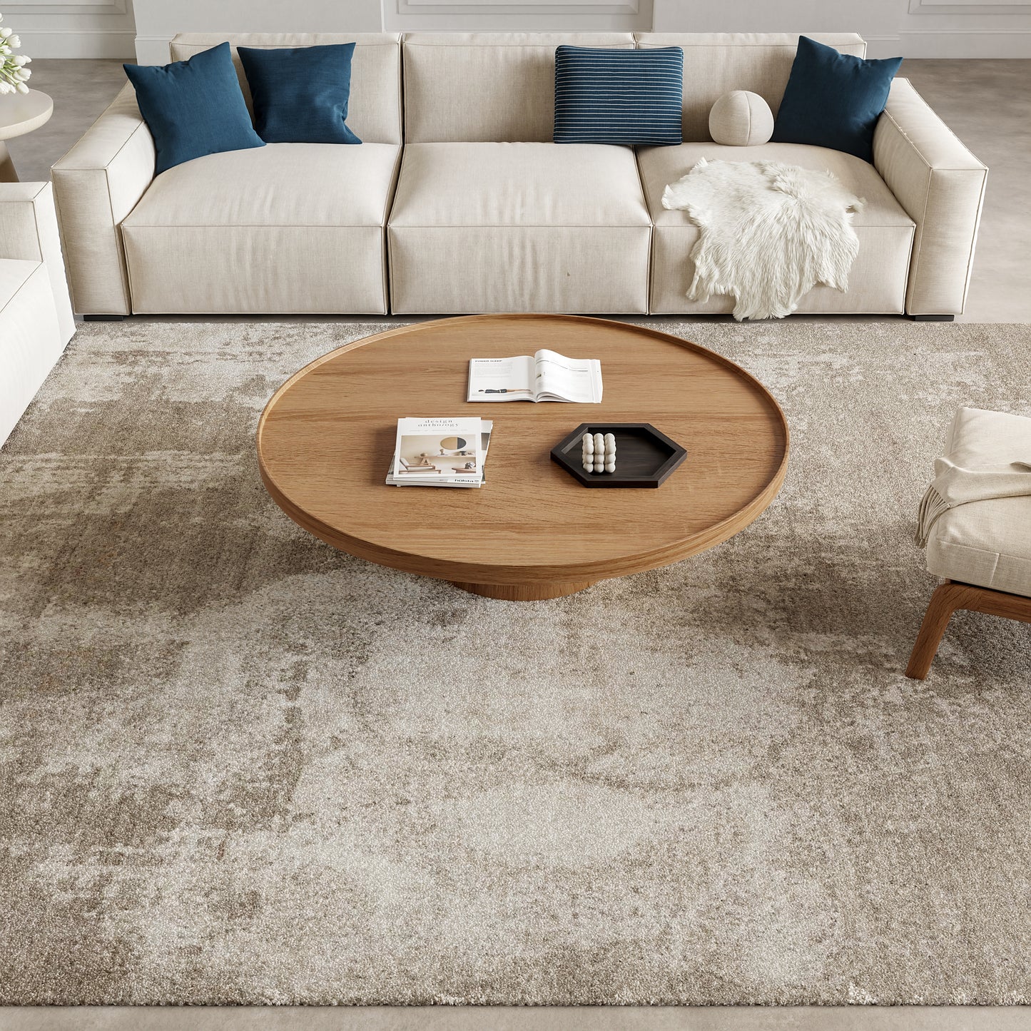 Rug under a round wooden coffee table in front of a beige sectional with blue pillows & a white throw; magazines on the table.