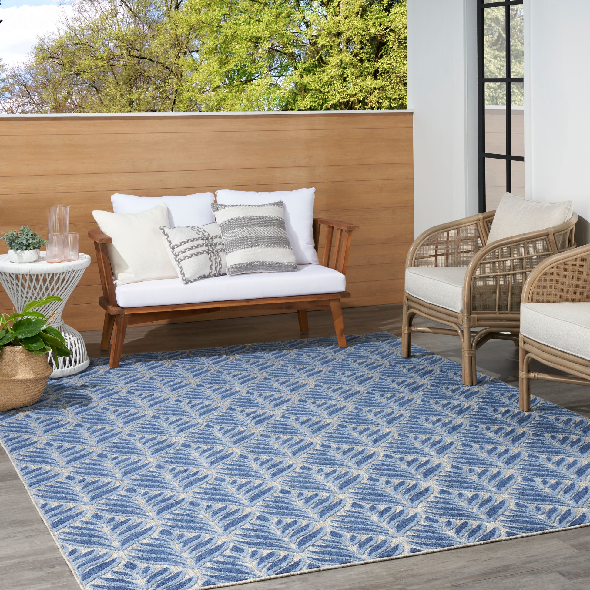 Blue outdoor patterned rug with leaf motif, white-cushioned wooden sofa and beige wicker chairs on a balcony with plants.