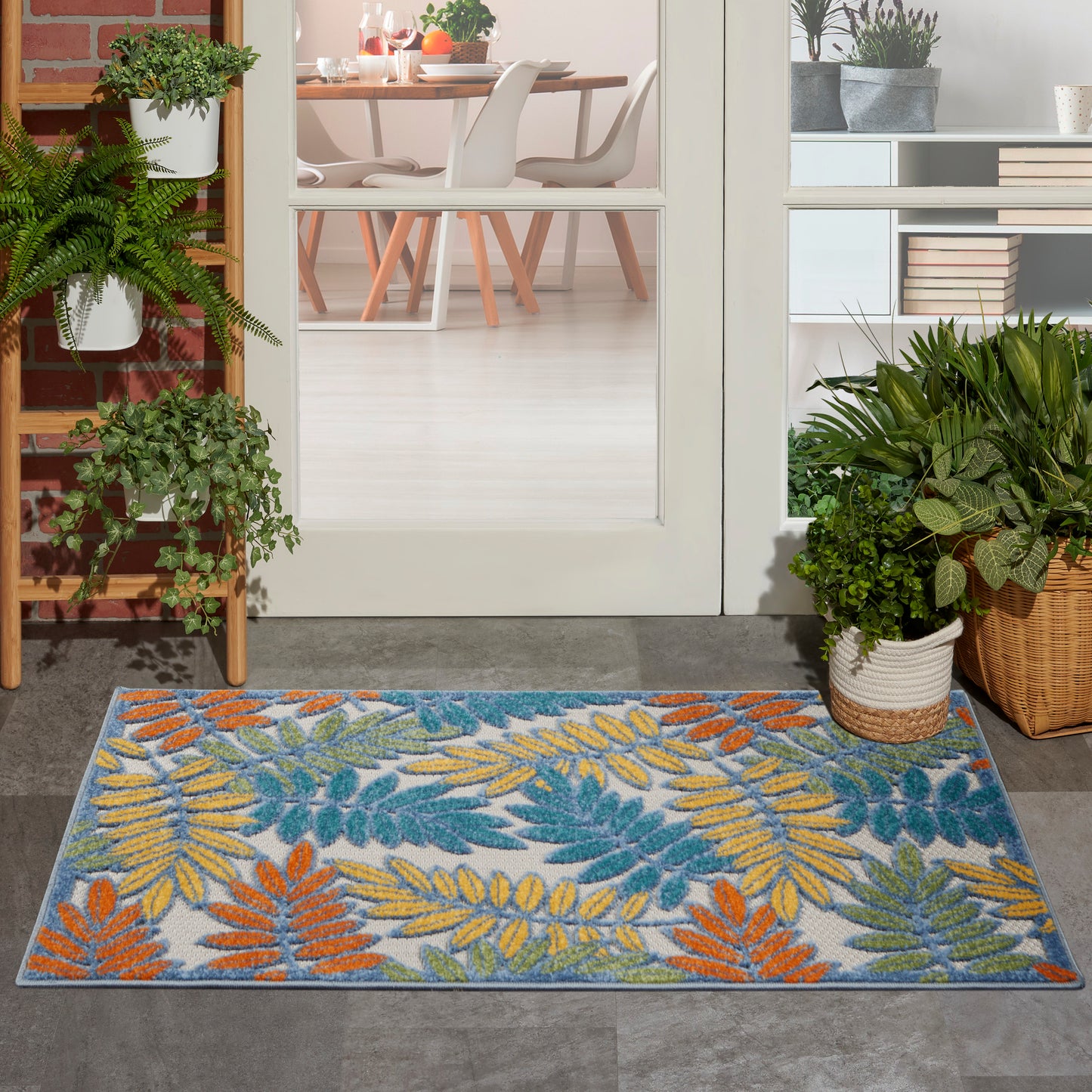 Colorful leaf-pattern outdoor doormat with blue-gray background and orange, yellow, and green leaves at a glass door threshold.