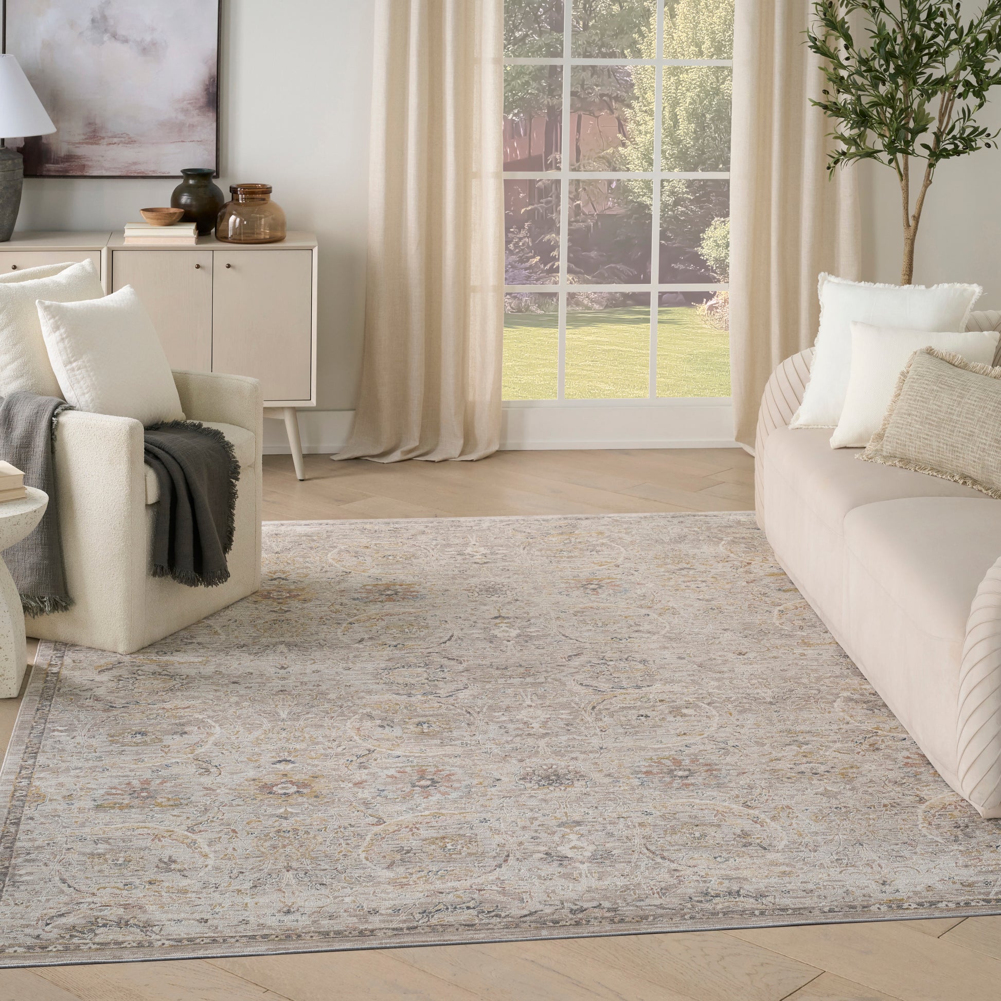 Neutral living room with large traditional-patterned area rug in gray-blue and beige, cream sofa, armchair, and tall plant.
